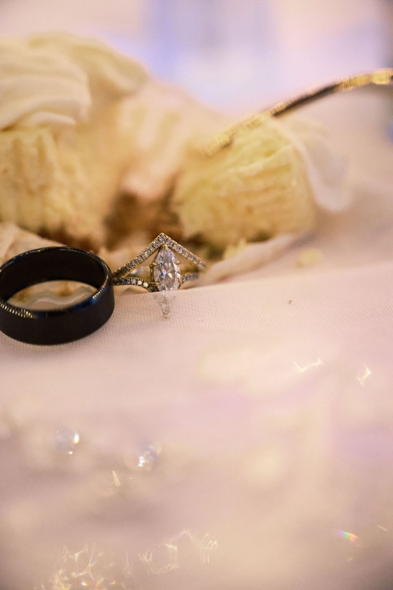 Close-up of a marquise diamond engagement ring beside a black wedding band on a white reflective surface.