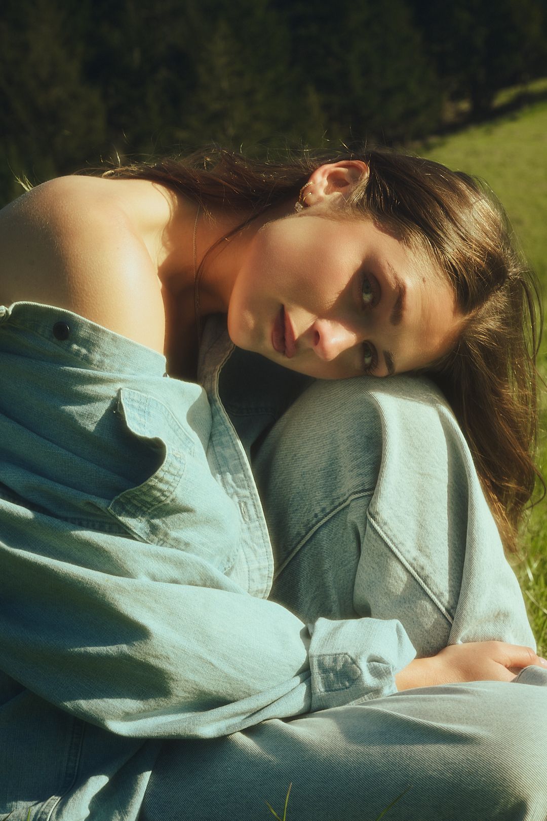 Young woman in denim shirt resting on grass in sunlight, looking at camera with serene expression outdoors.