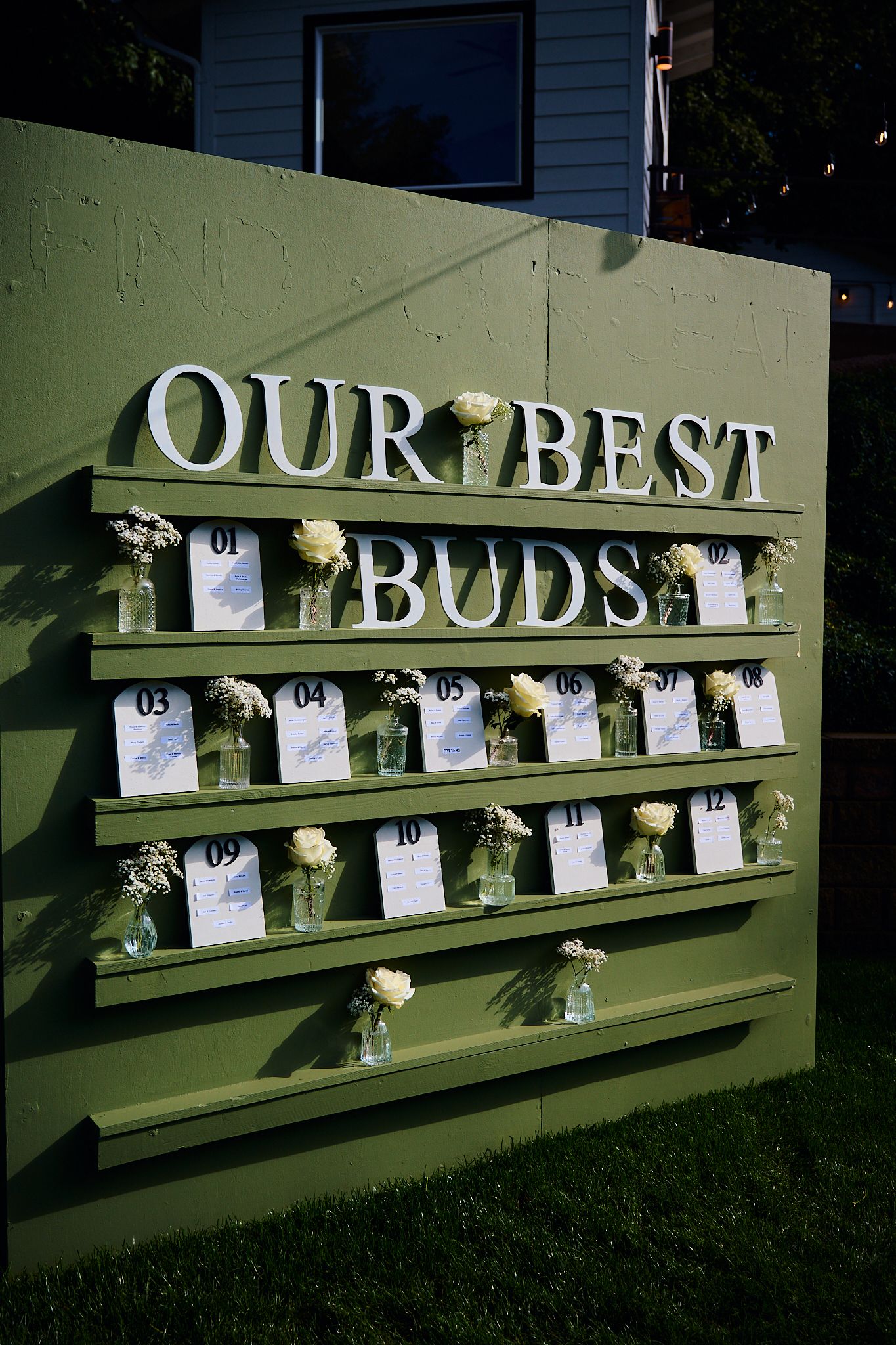 Green wedding seating chart display with “Our Best Buds” sign, numbered cards, and mason jar floral arrangements.