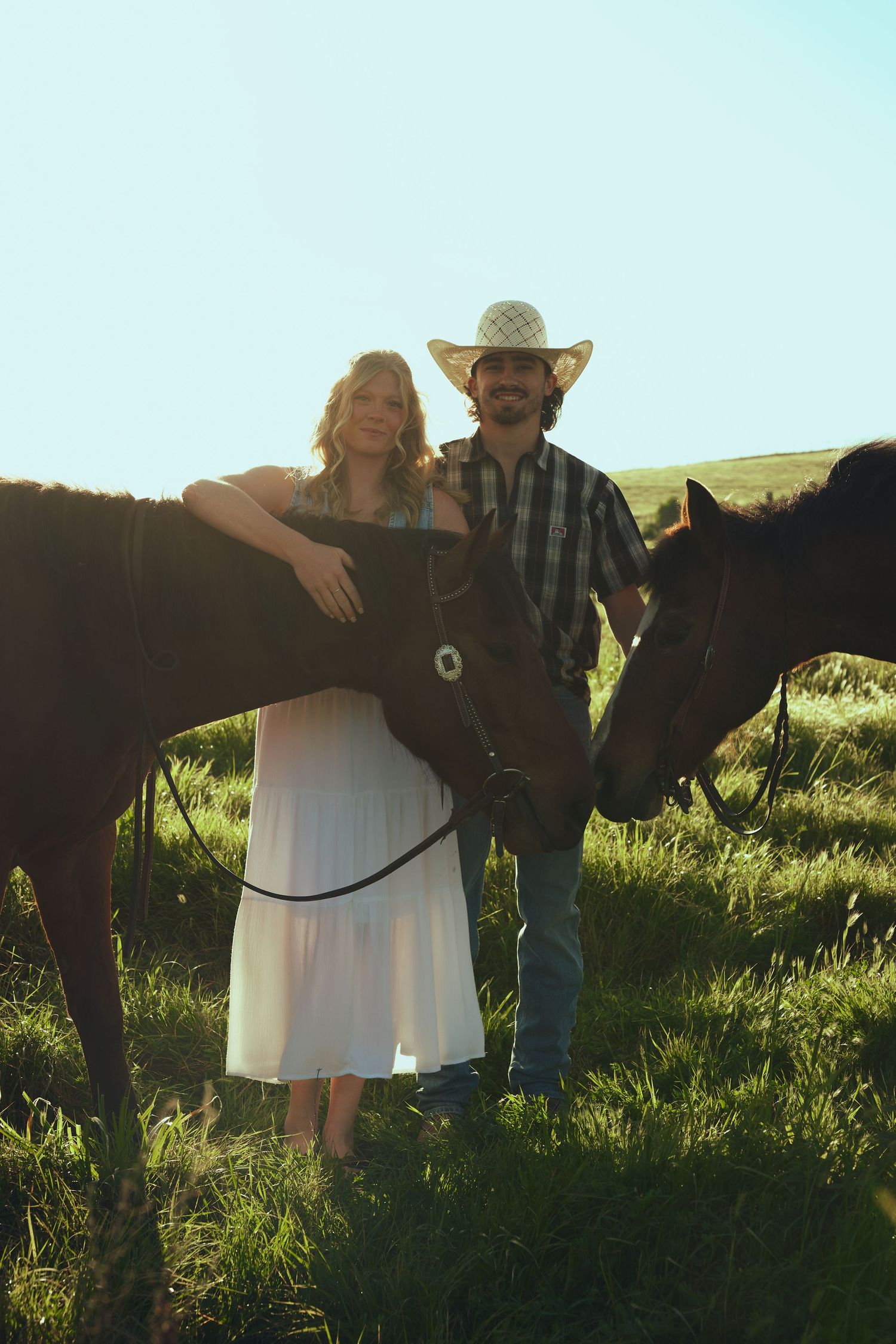 A couple stands with two horses in a sunlit green field, the man wearing a cowboy hat and plaid shirt.