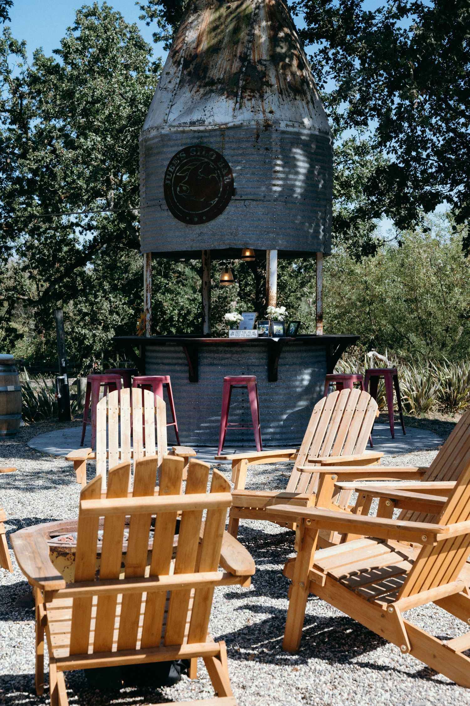 Wooden Adirondack chairs arranged on gravel patio facing rustic outdoor bar beneath vintage metal silo structure.