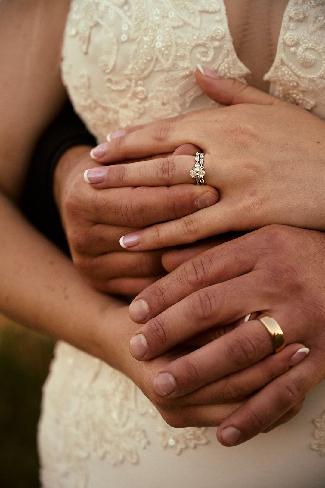 Bride and groom embracing, showing diamond engagement ring and gold wedding band on lace wedding gown.