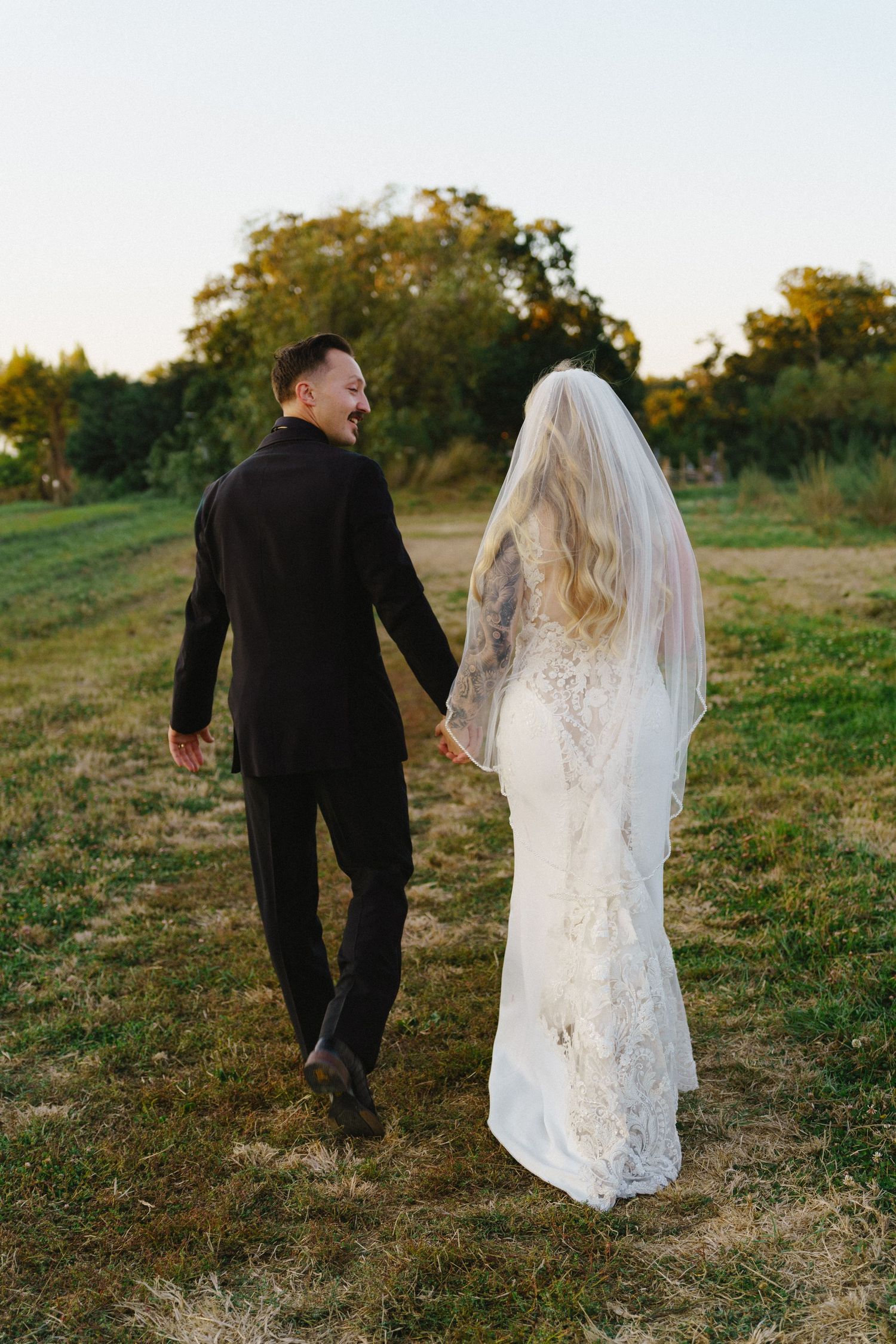 Couple in wedding attire walking through a grassy field at golden hour.