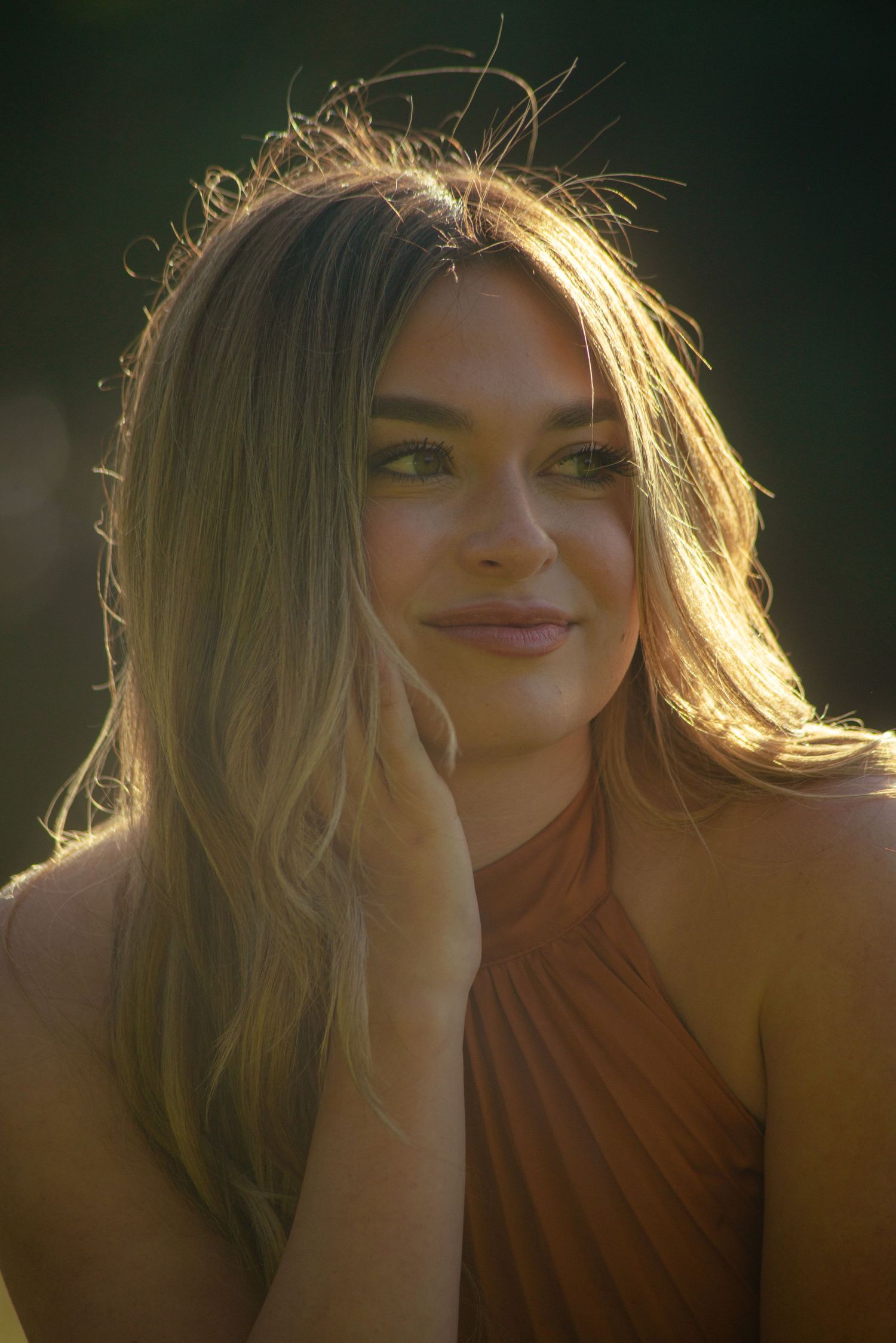 Portrait in warm lighting shows someone in a rust-colored halter dress against a dark background.
