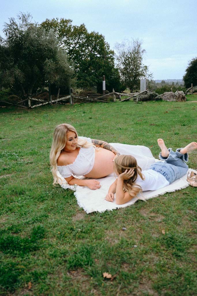 A pregnant woman and child rest on a white blanket in a pastoral setting with wooden fencing.