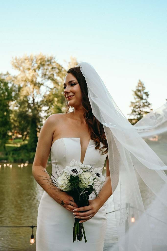 A bride in a strapless white dress holds a bouquet of white flowers by a lake at sunset.