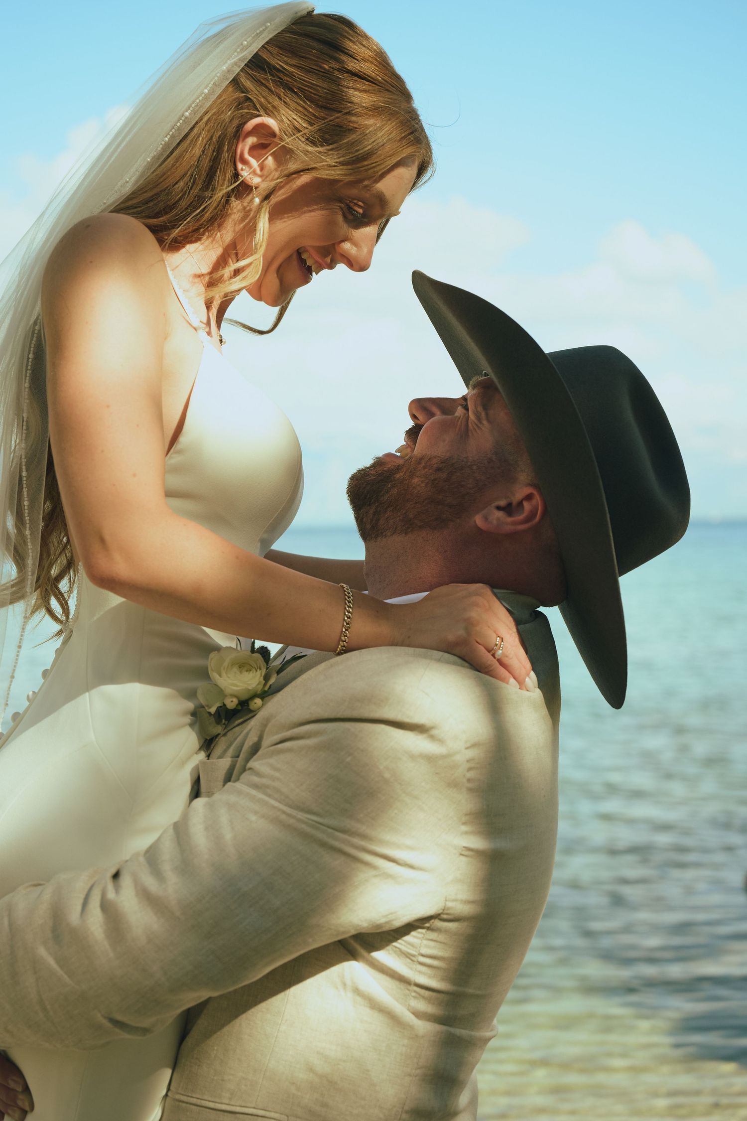 Bride and groom share a joyful moment by the ocean, groom lifting bride in a tan suit and black cowboy hat.