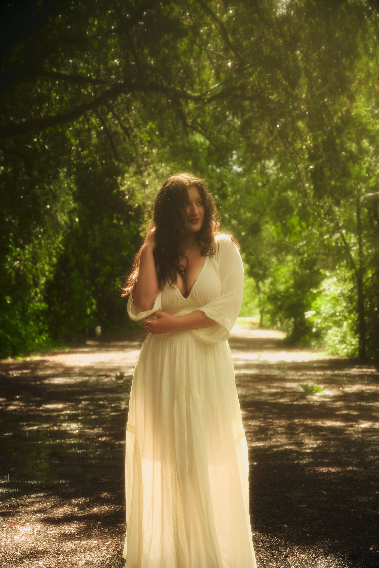 Woman in white flowing dress standing on a sunlit forest path surrounded by lush green trees.