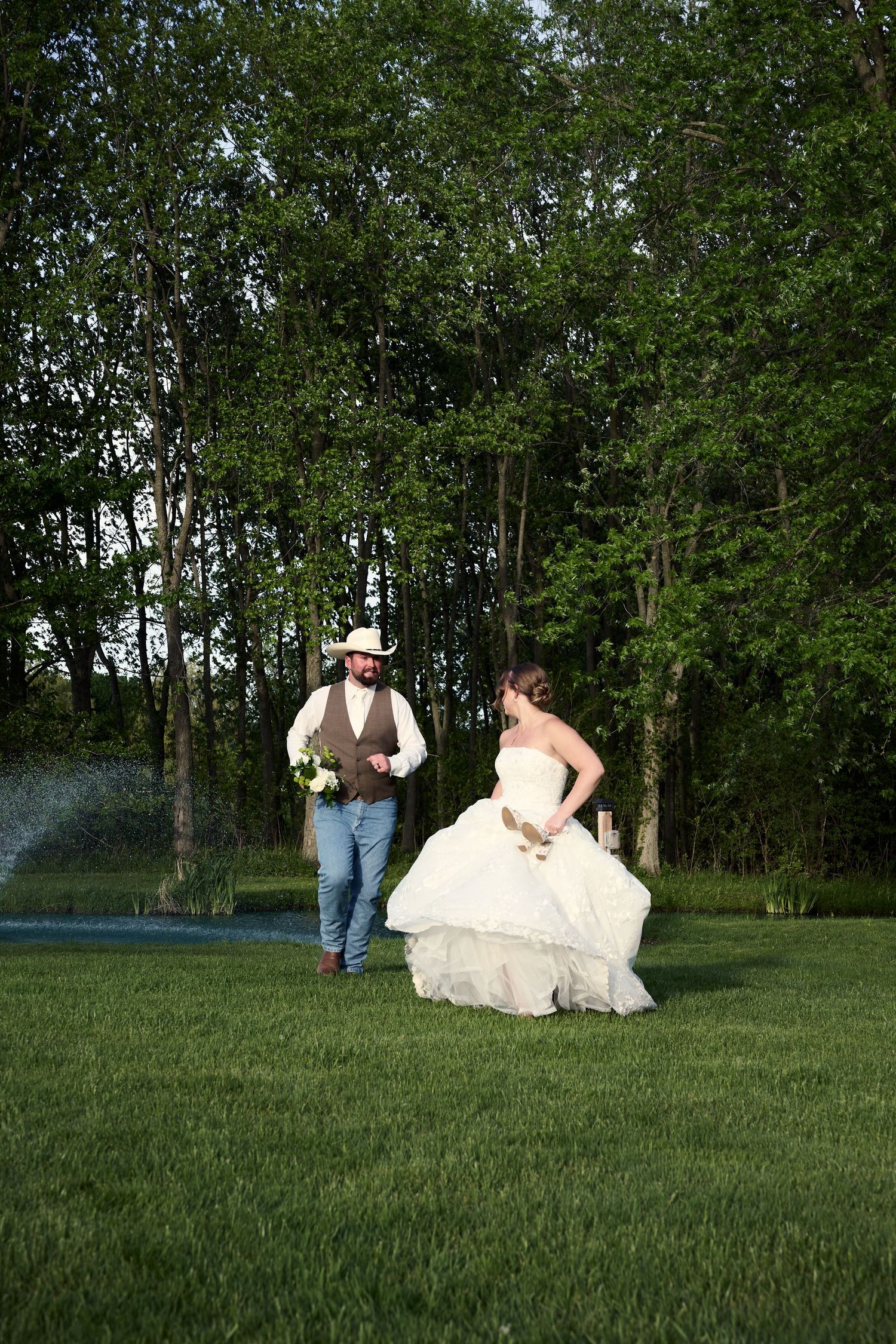 Bride in white gown and groom in cowboy hat walk together on green lawn with trees in background at outdoor wedding.
