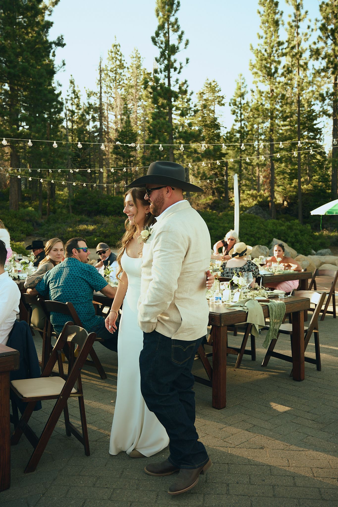 Bride and groom share first dance at outdoor rustic wedding reception surrounded by pine trees and string lights.
