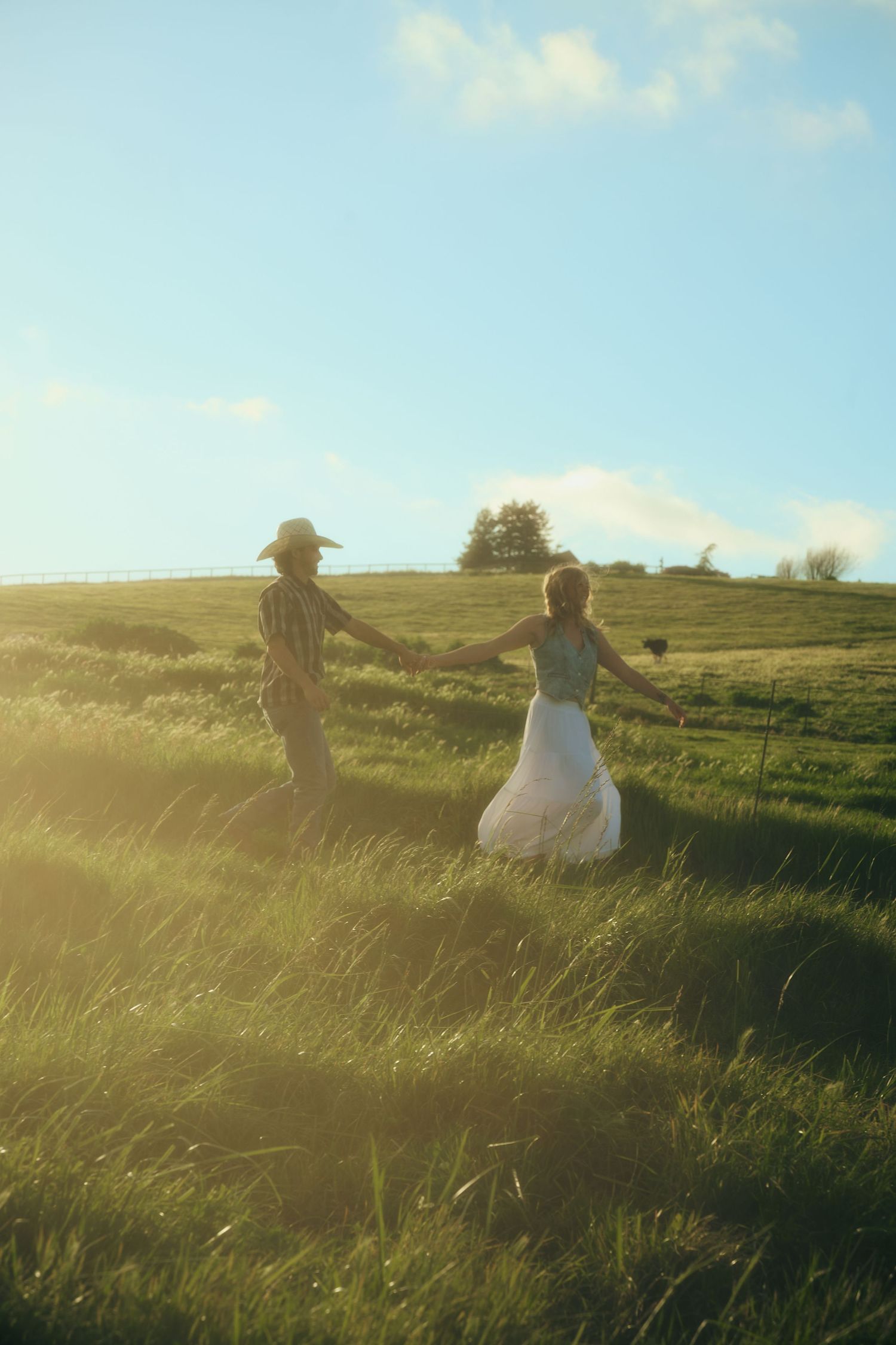 Couple holding hands walking through a sunlit green meadow, woman in white dress and man in cowboy hat.