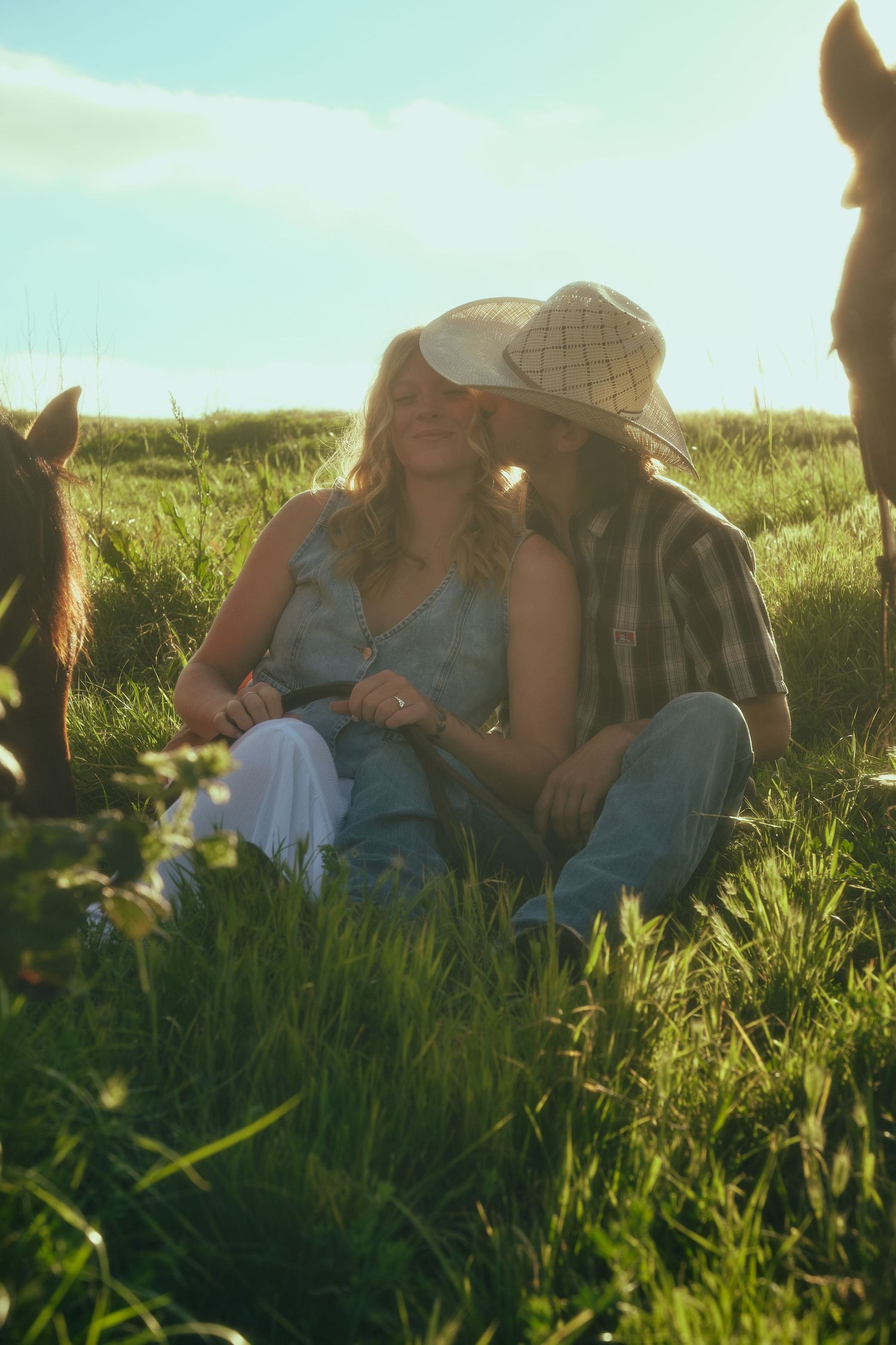 Romantic couple sitting in green field with horses, man in cowboy hat kissing woman in golden sunlight.