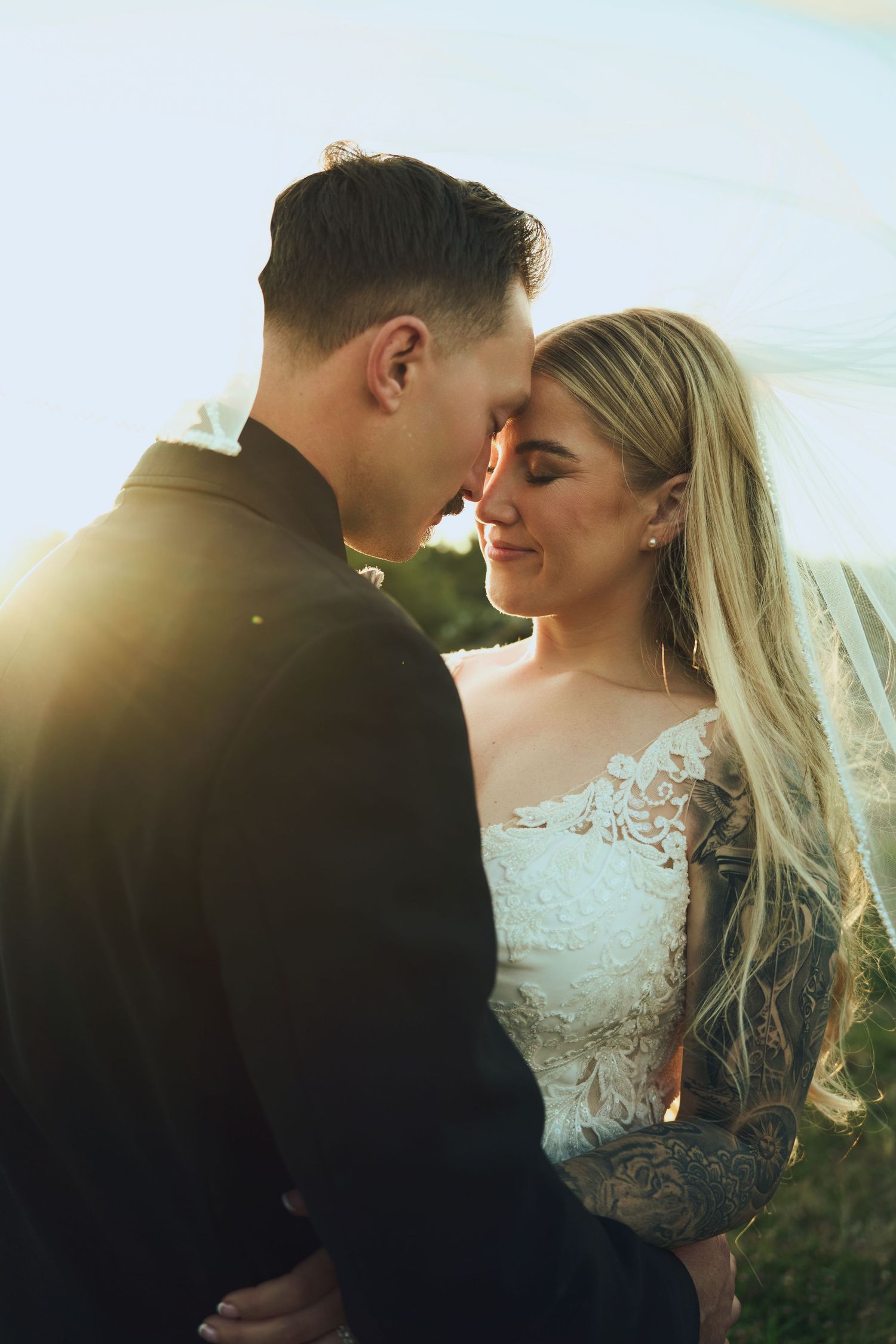 Bride and groom touch foreheads romantically in golden sunset light, bride wearing lace wedding gown.