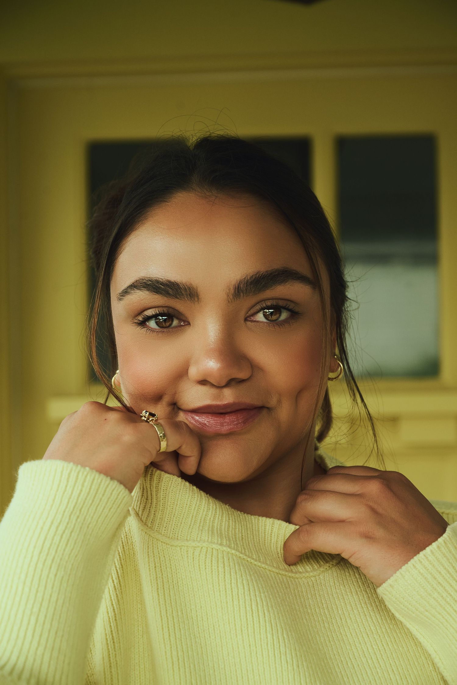 Young woman in yellow sweater smiling warmly, holding collar with ringed fingers, green eyes, natural makeup.