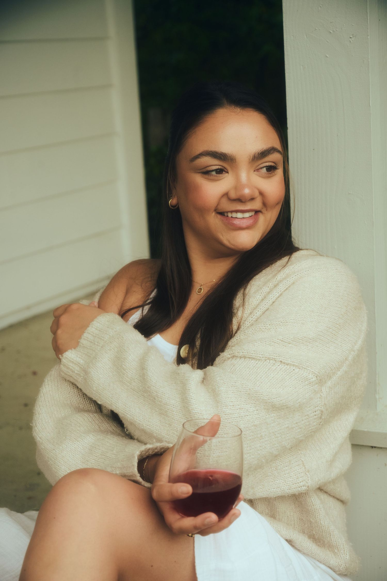 Smiling young woman in a beige cardigan holding a stemless glass of red wine while sitting on a porch.