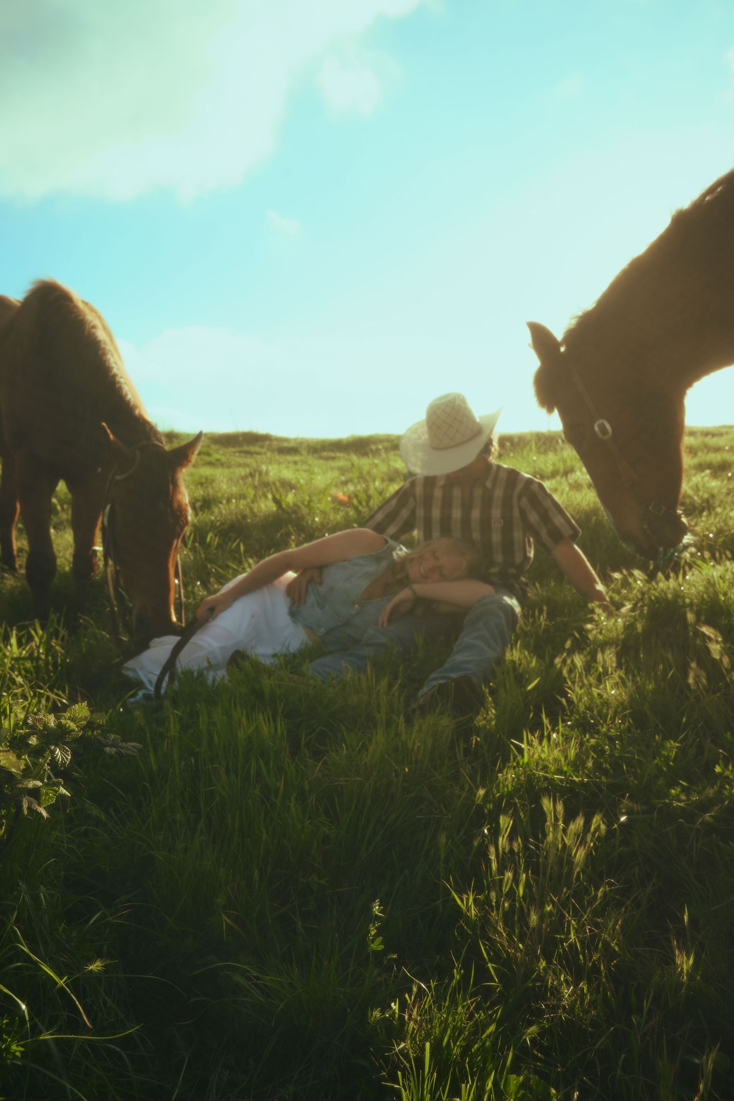 Couple relaxing in green pasture wearing cowboy hat, surrounded by grazing brown horses under blue sky.