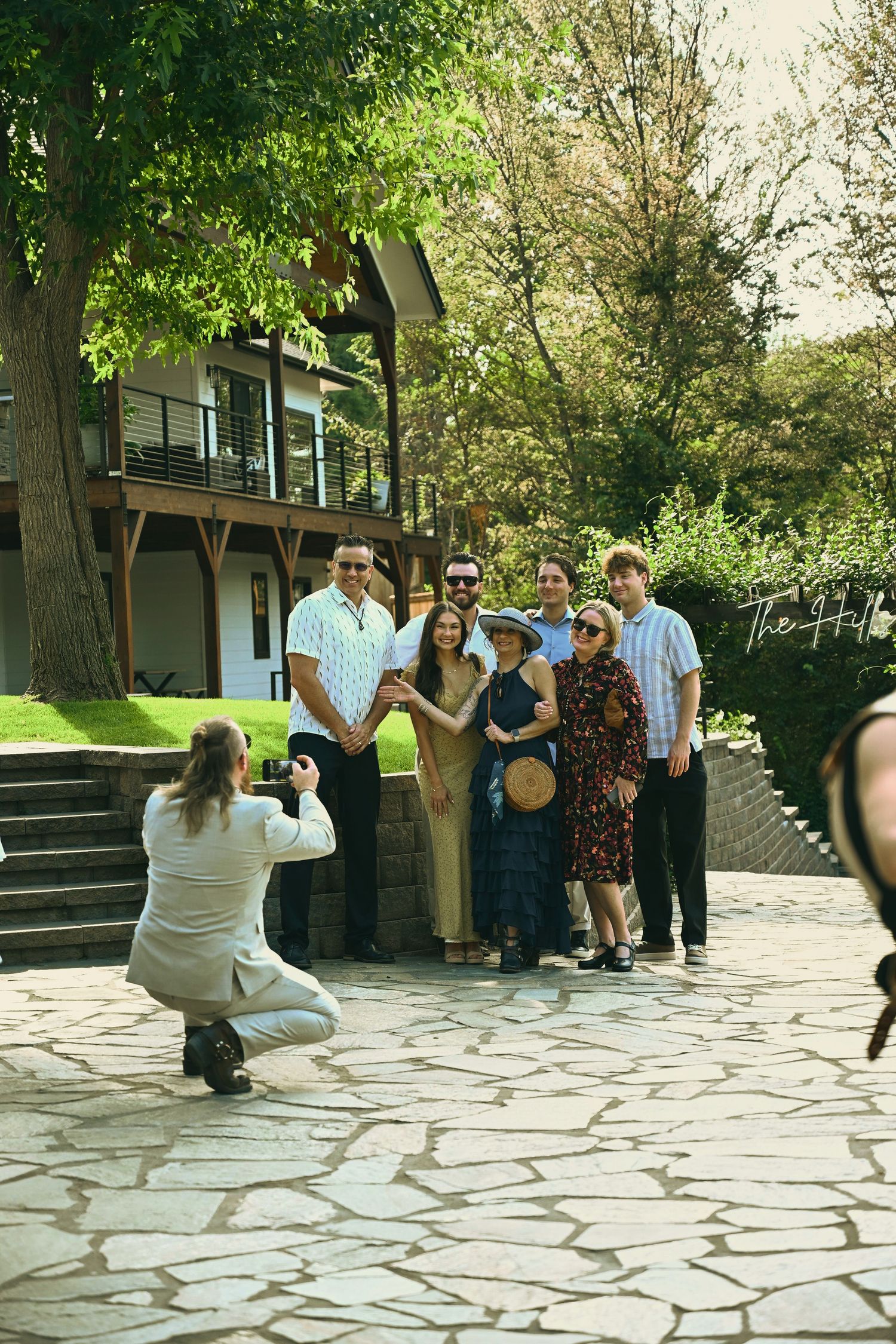 Photographer crouching to capture a group of six people posing outdoors near a stone patio and lush garden.