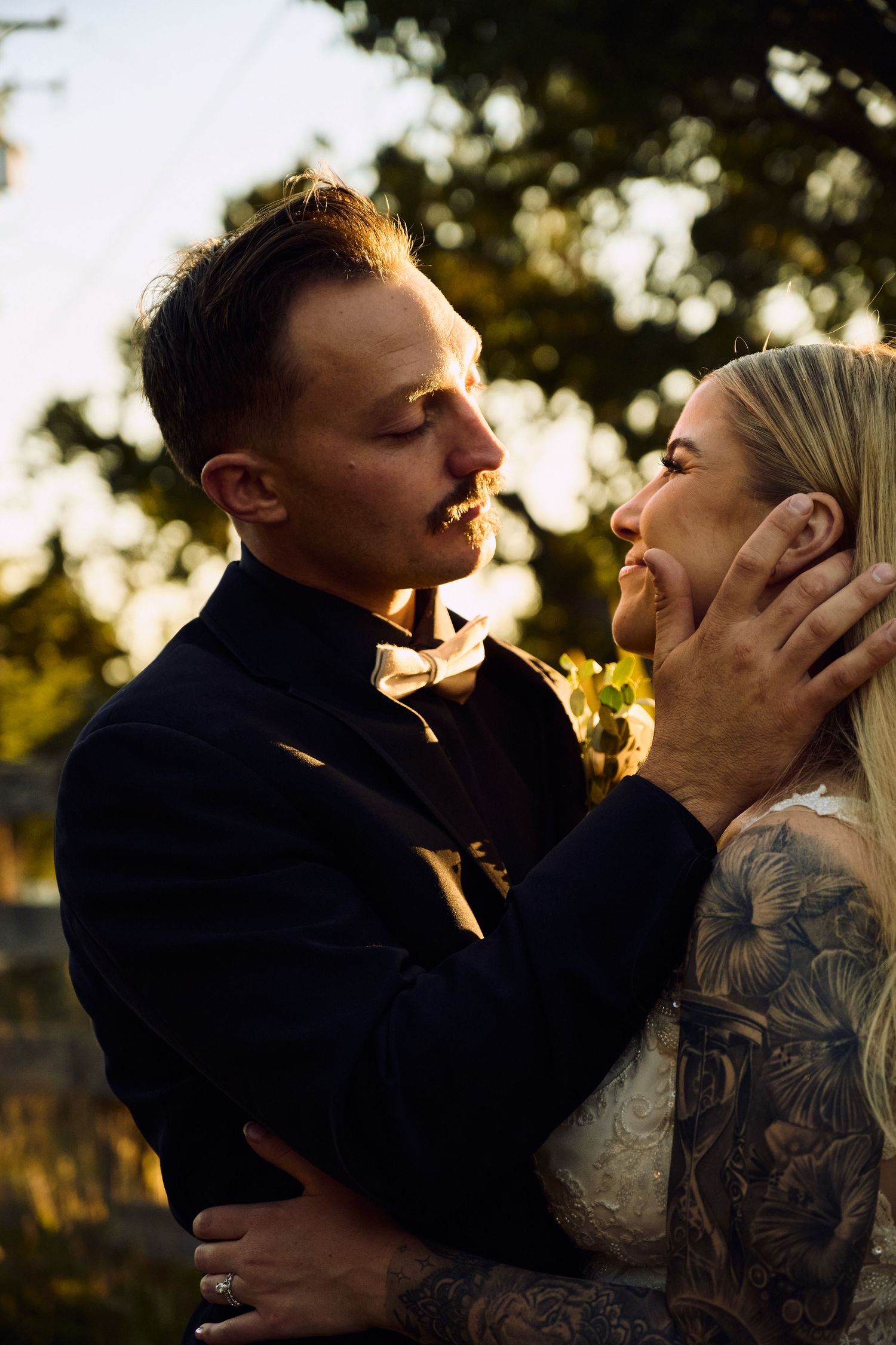 Tattooed couple in wedding attire share an intimate moment at golden hour, groom tenderly holding bride's face.