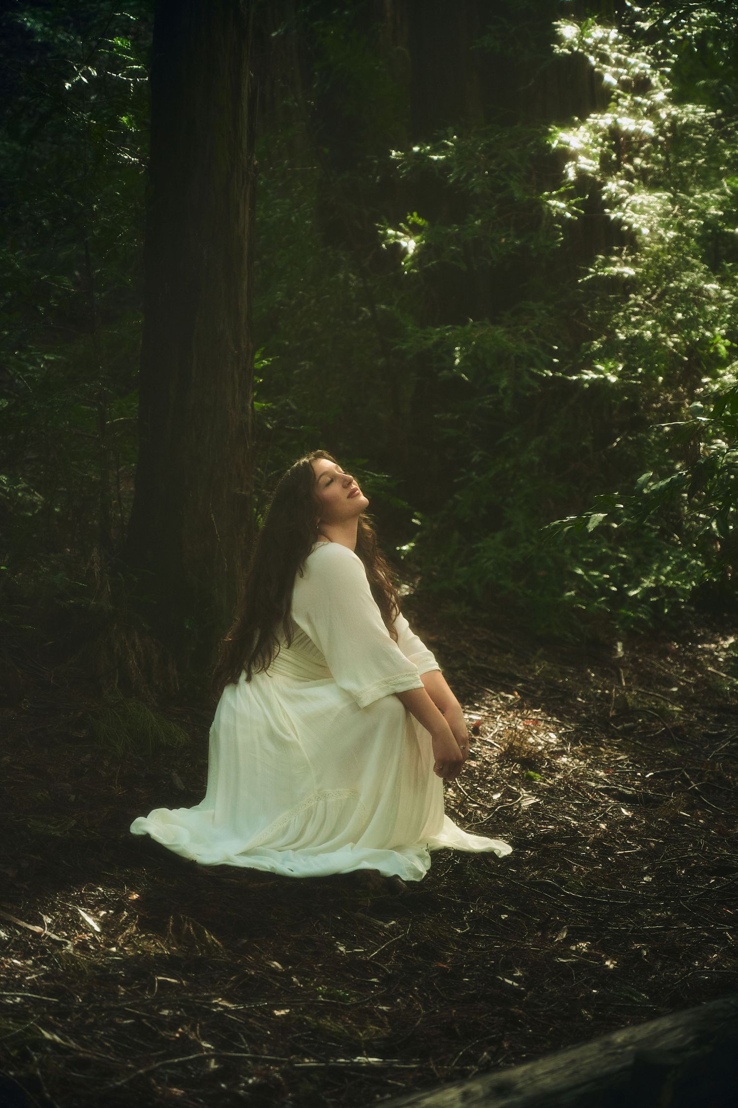 Woman in white dress sits on log in dark forest, bathed in dappled sunlight filtering through tall trees.