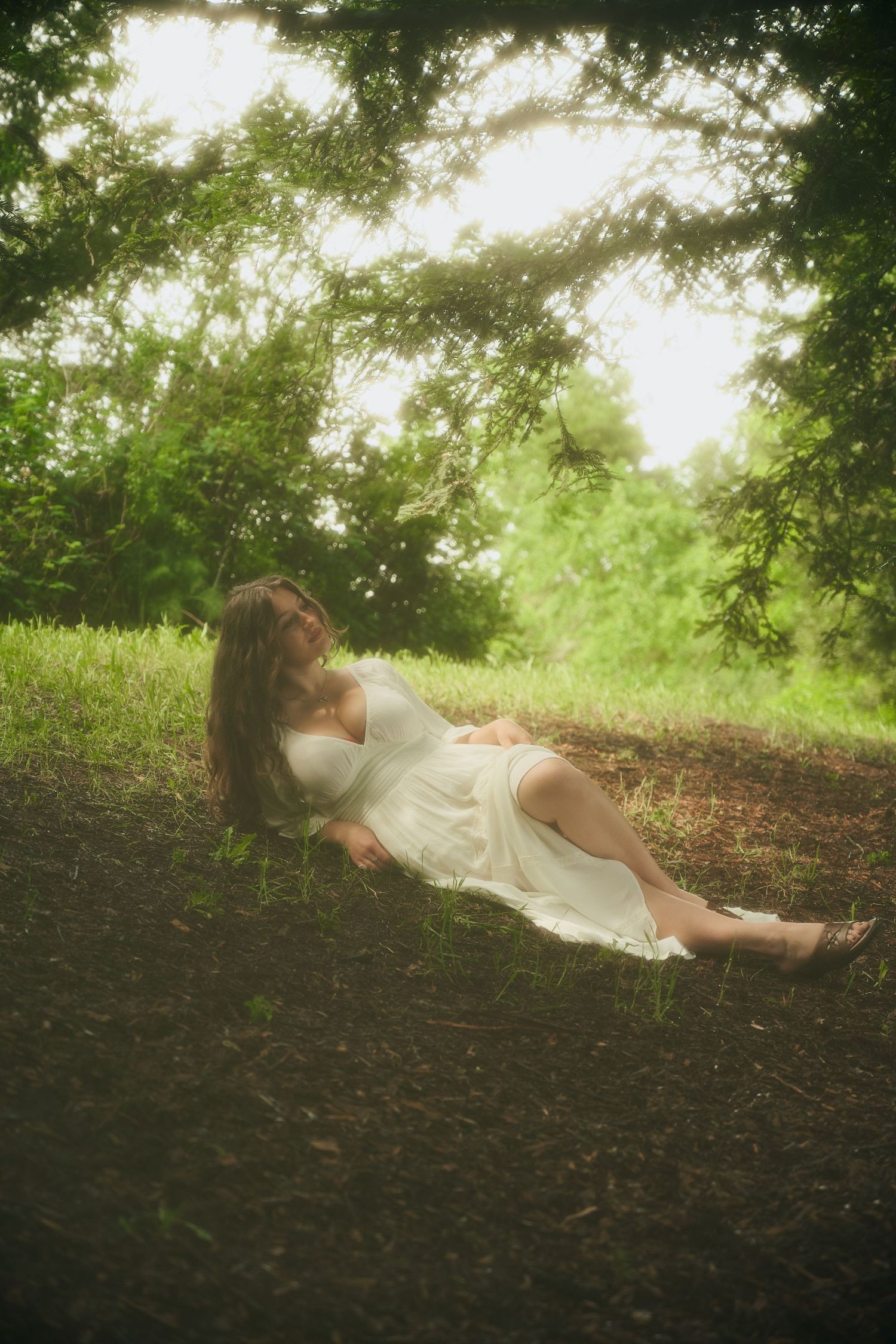 Woman in white dress reclining peacefully under lush green trees in a dreamy, softly lit forest setting.