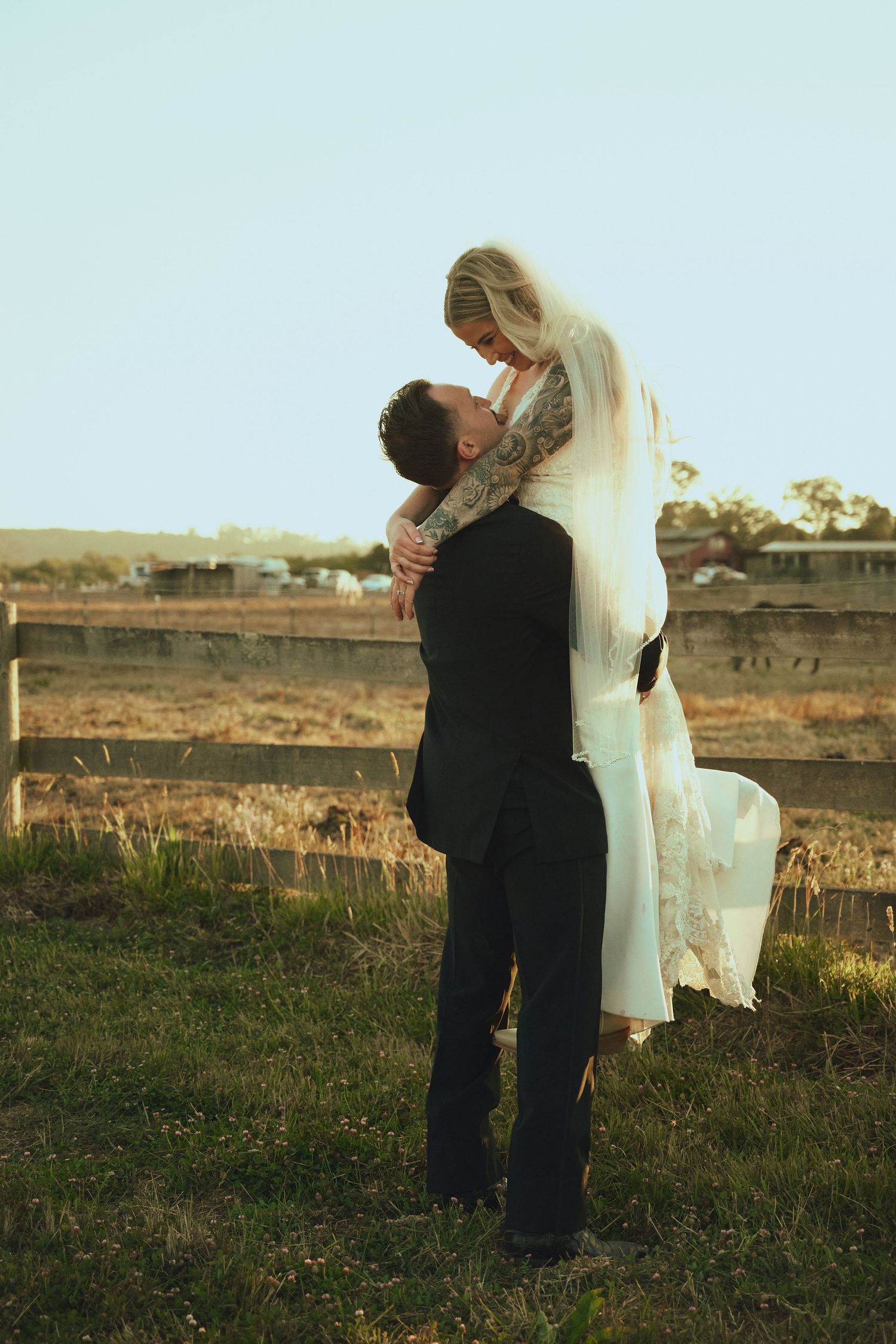 Groom lifts bride in white lace gown and veil at sunset near a rustic fence in a countryside setting.