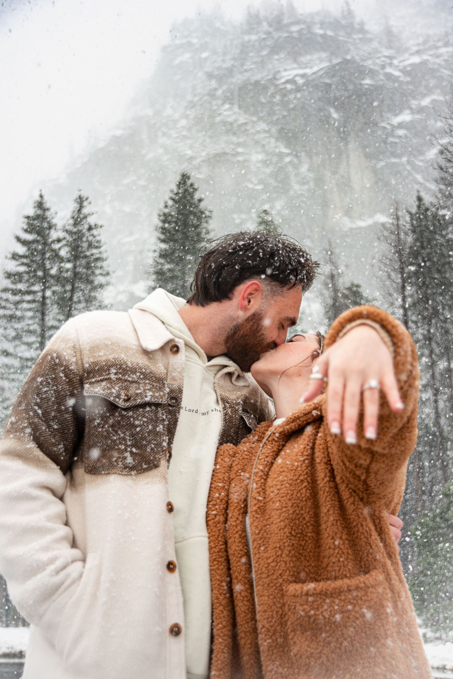 A couple embraces against a snowy mountain backdrop with evergreen trees.