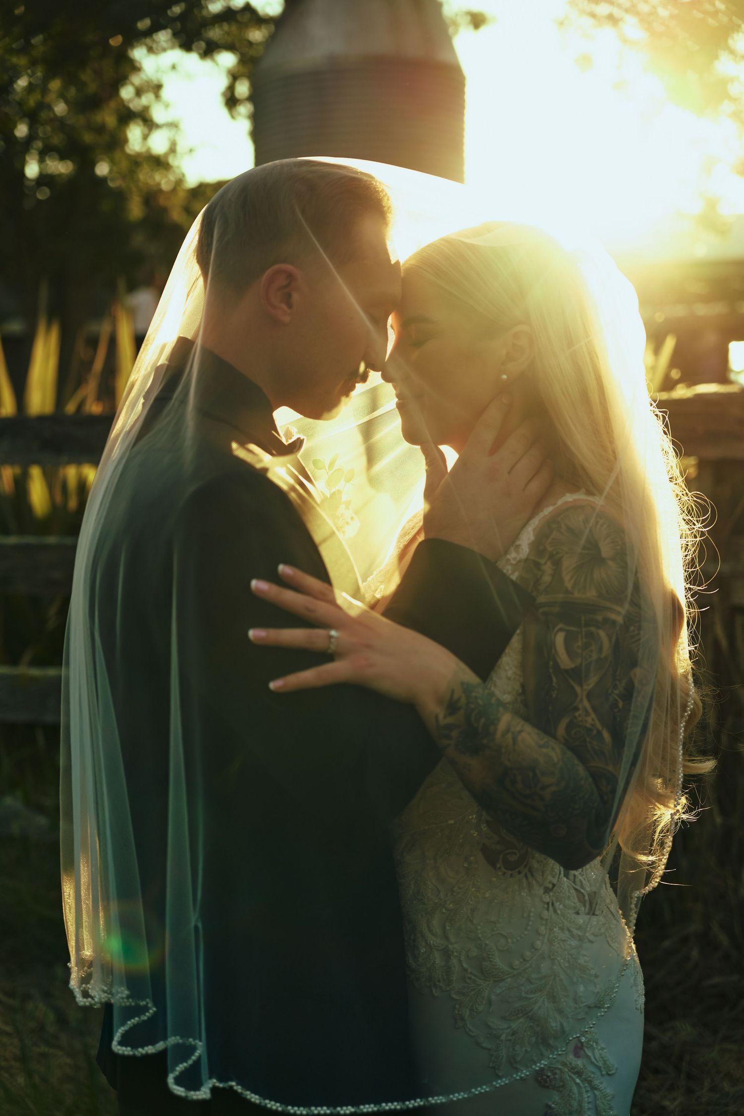 Bride and groom sharing a romantic moment under a veil with golden sunset backlight at outdoor wedding.