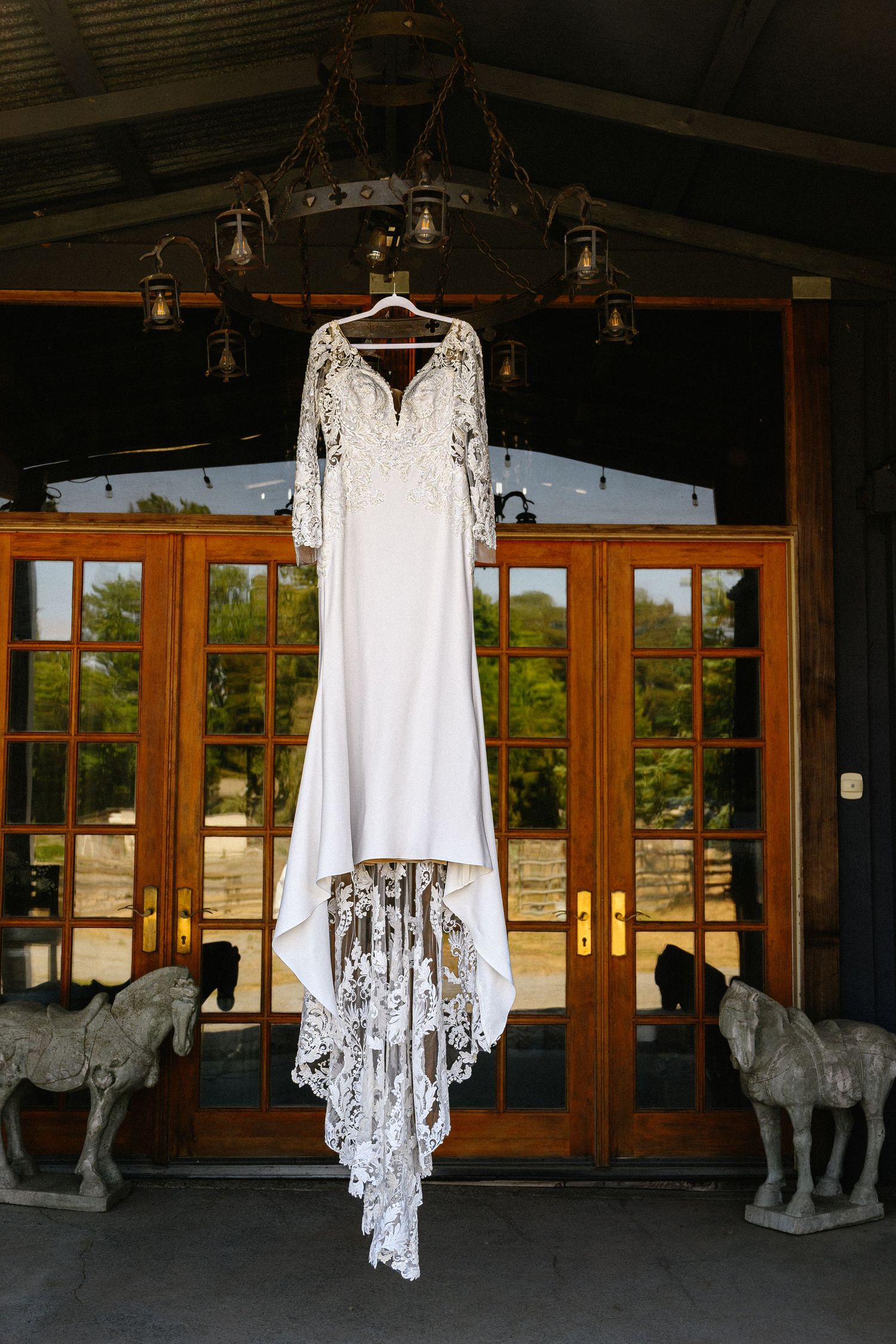 Elegant lace wedding dress with long sleeves hanging from a chandelier in a rustic venue with French doors.