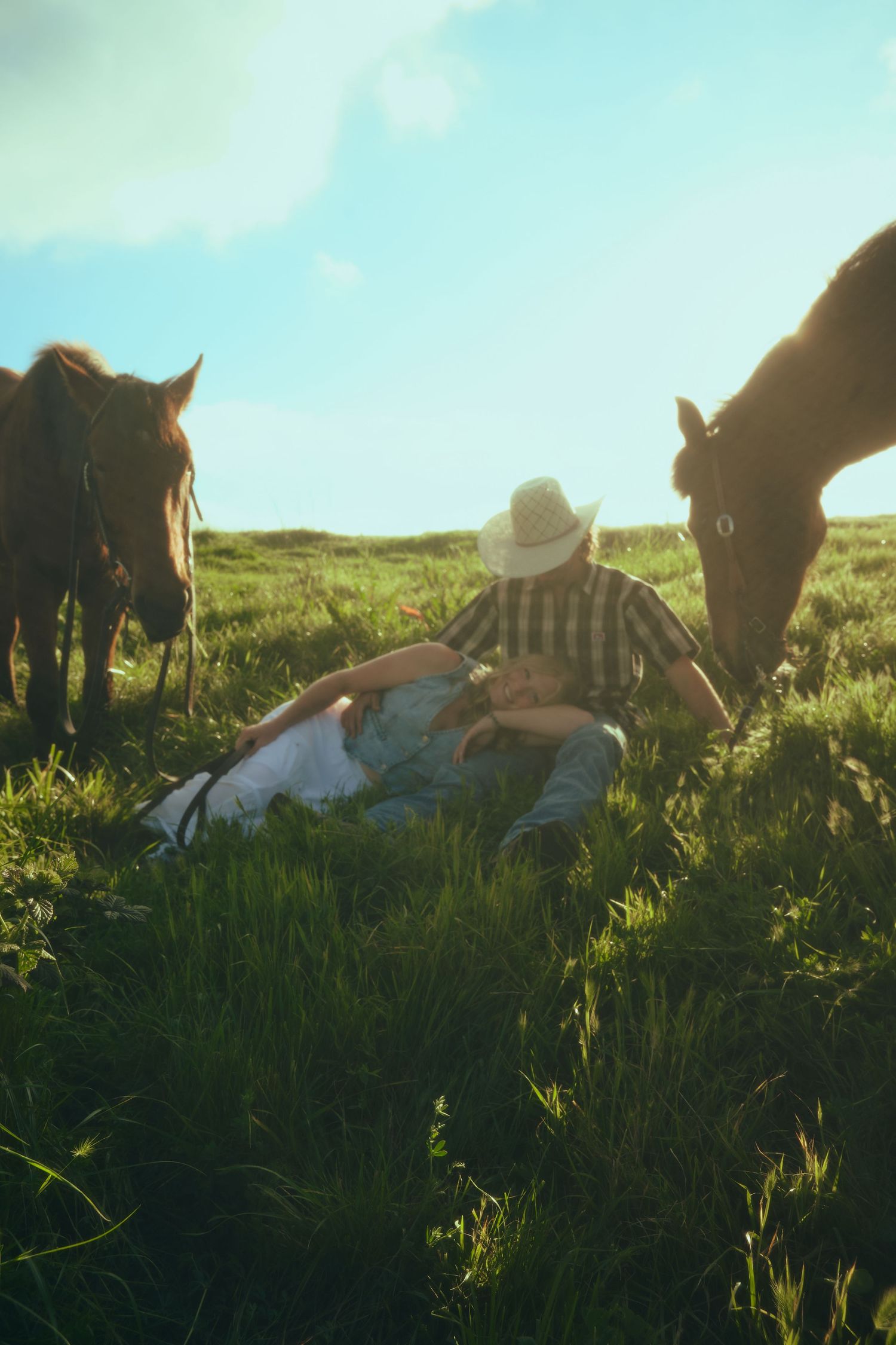 Couple sitting in green pasture with horses grazing nearby under a bright blue sky on a sunny day.