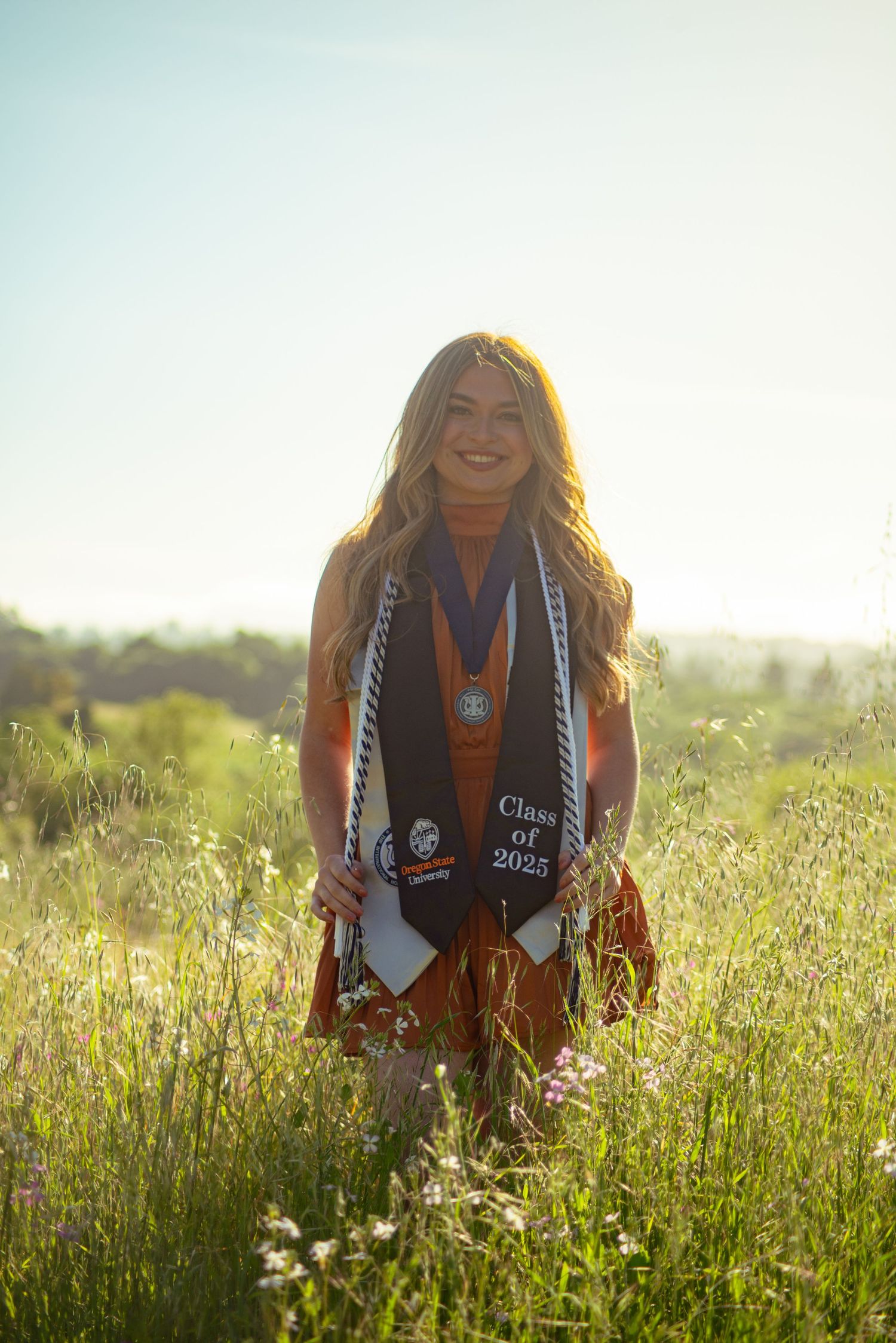 A graduate wearing academic regalia poses in a sunlit meadow with wildflowers.