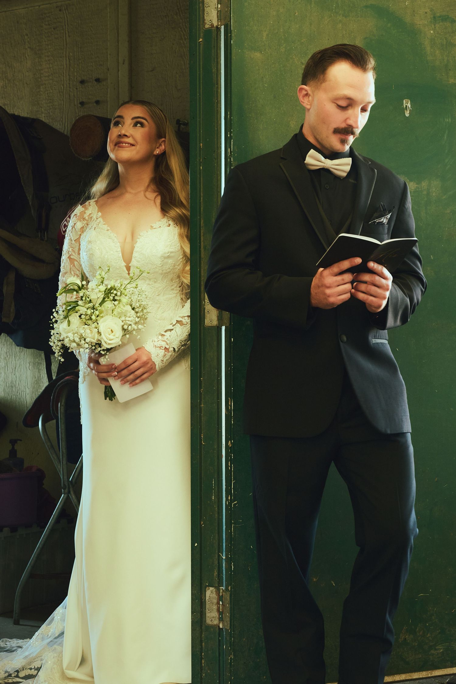 Bride in lace gown holding bouquet stands beside groom in black tuxedo reading vows before wedding ceremony.