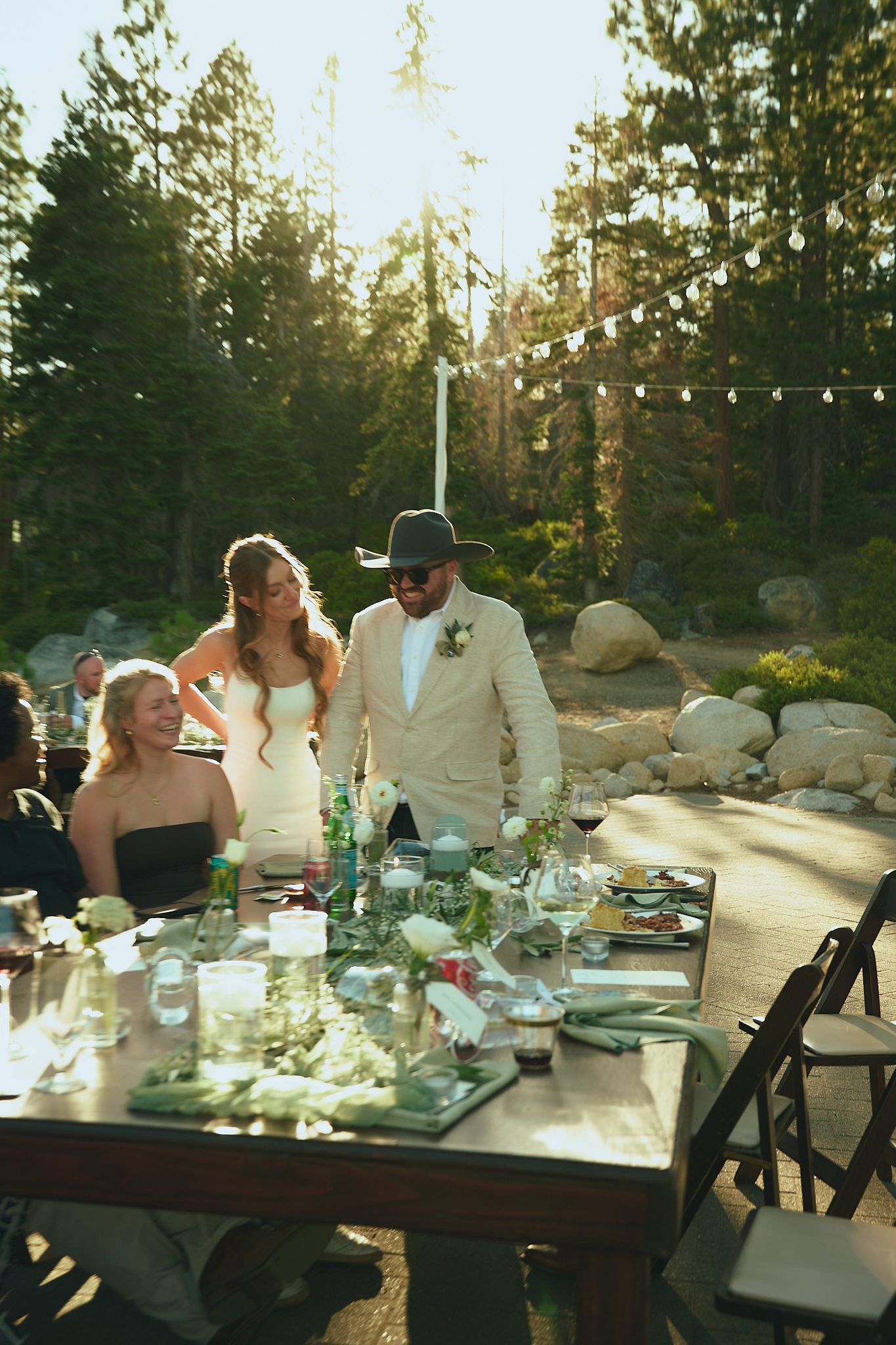 Bride and groom standing together at outdoor wedding reception table surrounded by pine trees and string lights.