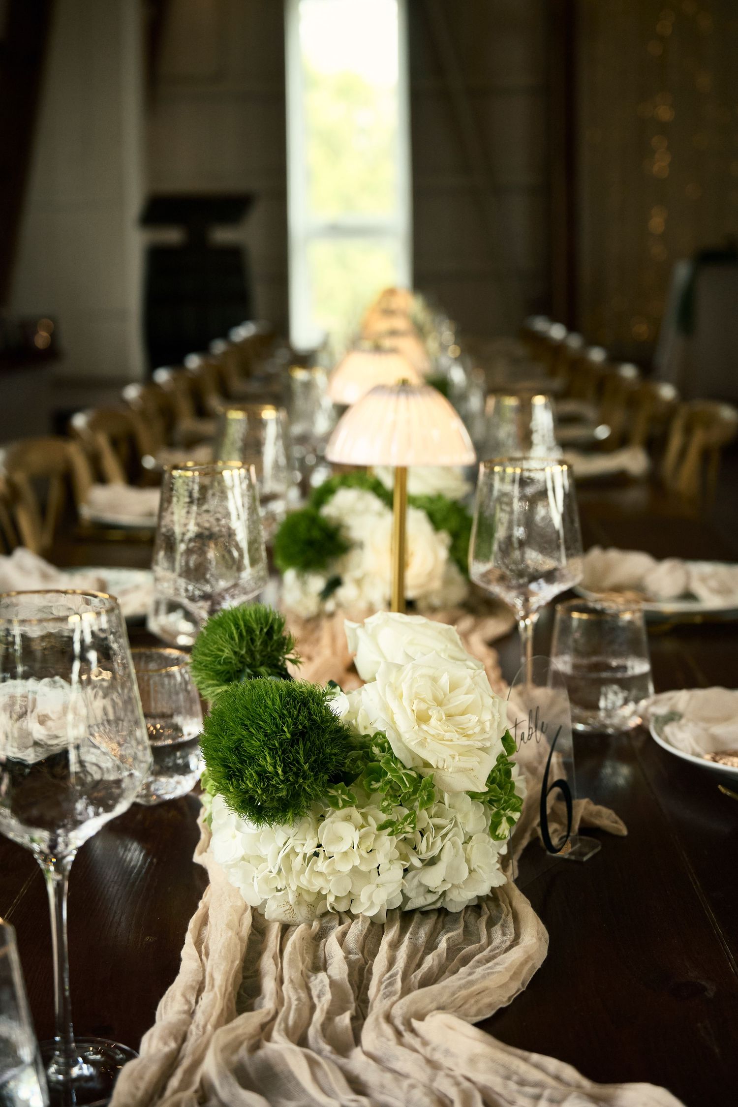 Elegant wedding reception table with white roses, green flowers, soft draping, crystal glasses, and a small lamp centerpiece.
