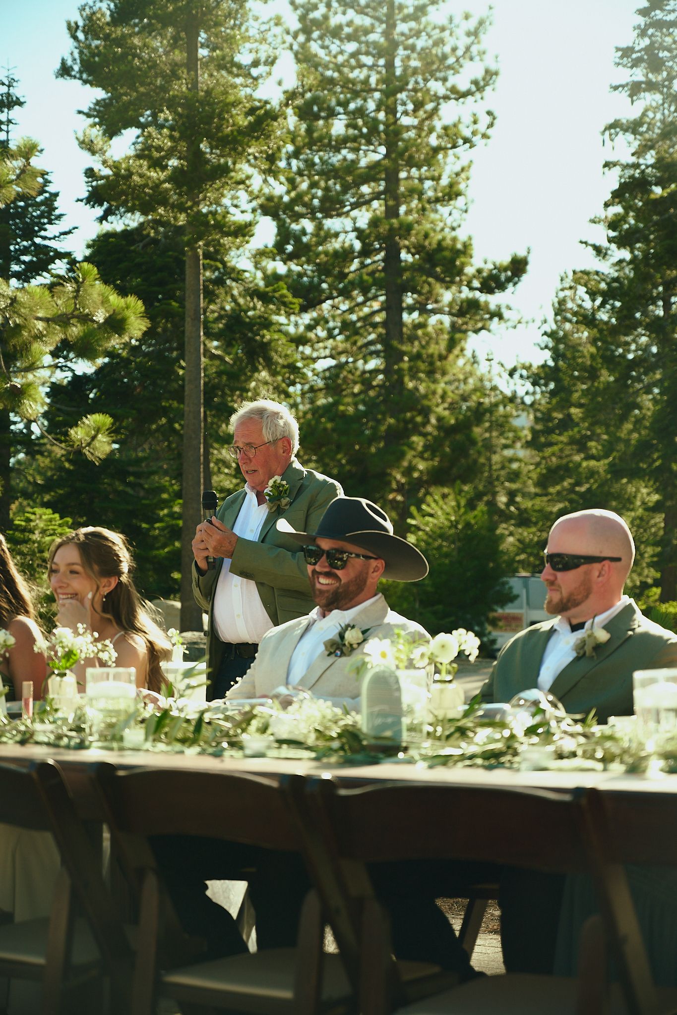 Outdoor wedding reception with guests seated at a decorated table, surrounded by tall pine trees on a sunny day.