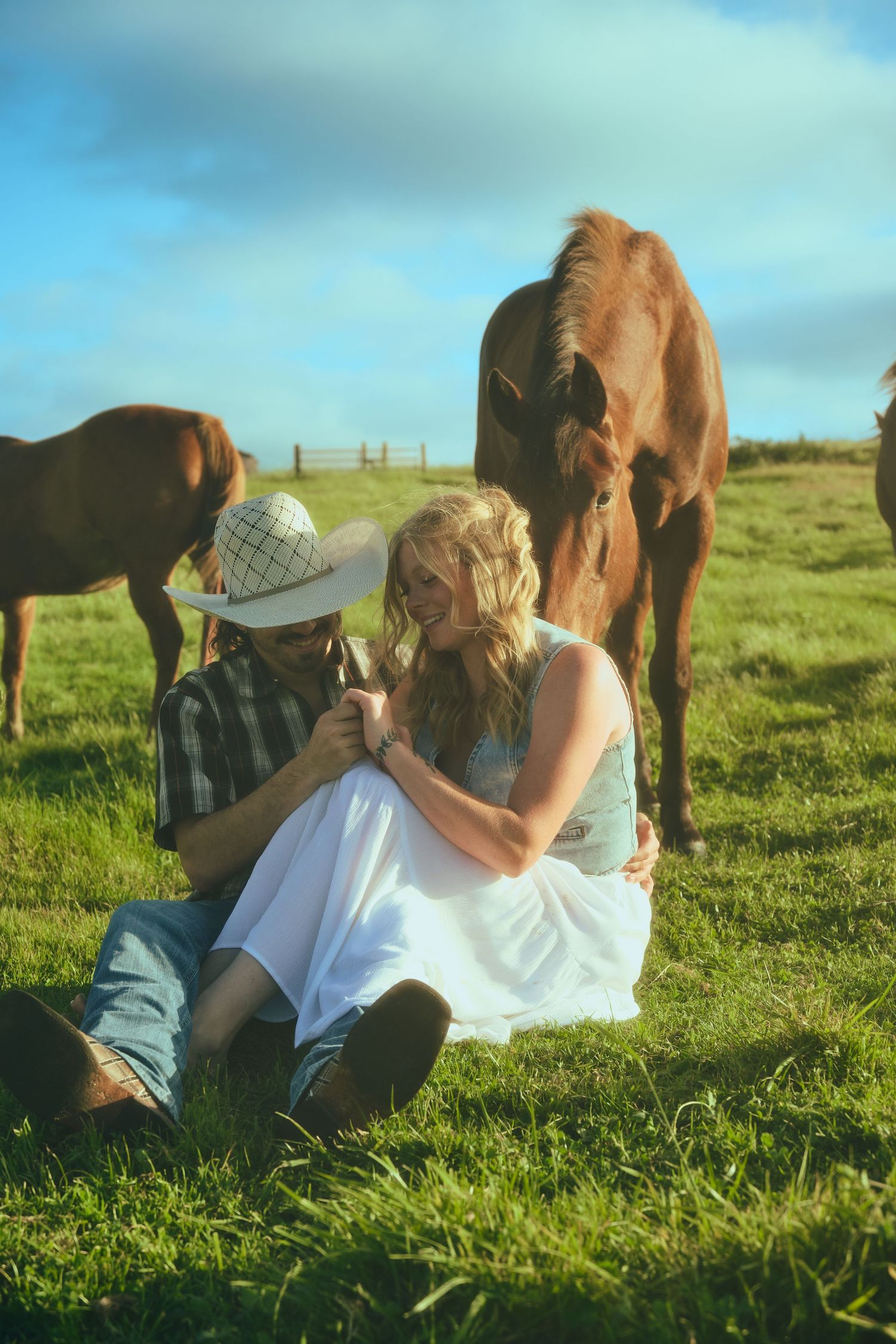 Couple sitting on green grass with brown horses grazing behind them on a sunny farm day.