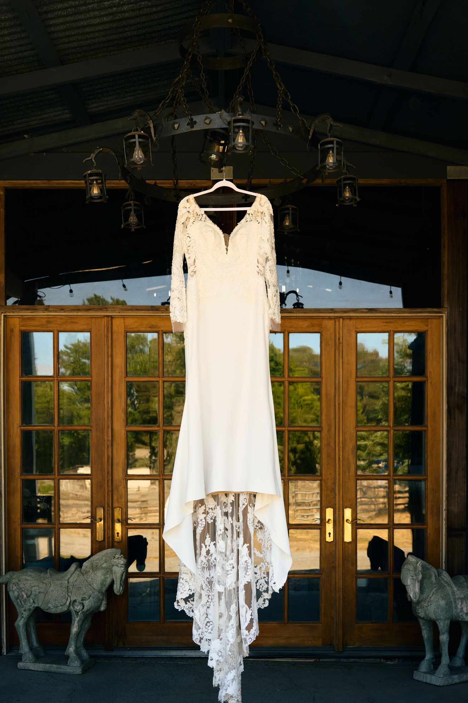 White lace wedding dress with long sleeves hanging from rustic chandelier above French doors with horse statues.