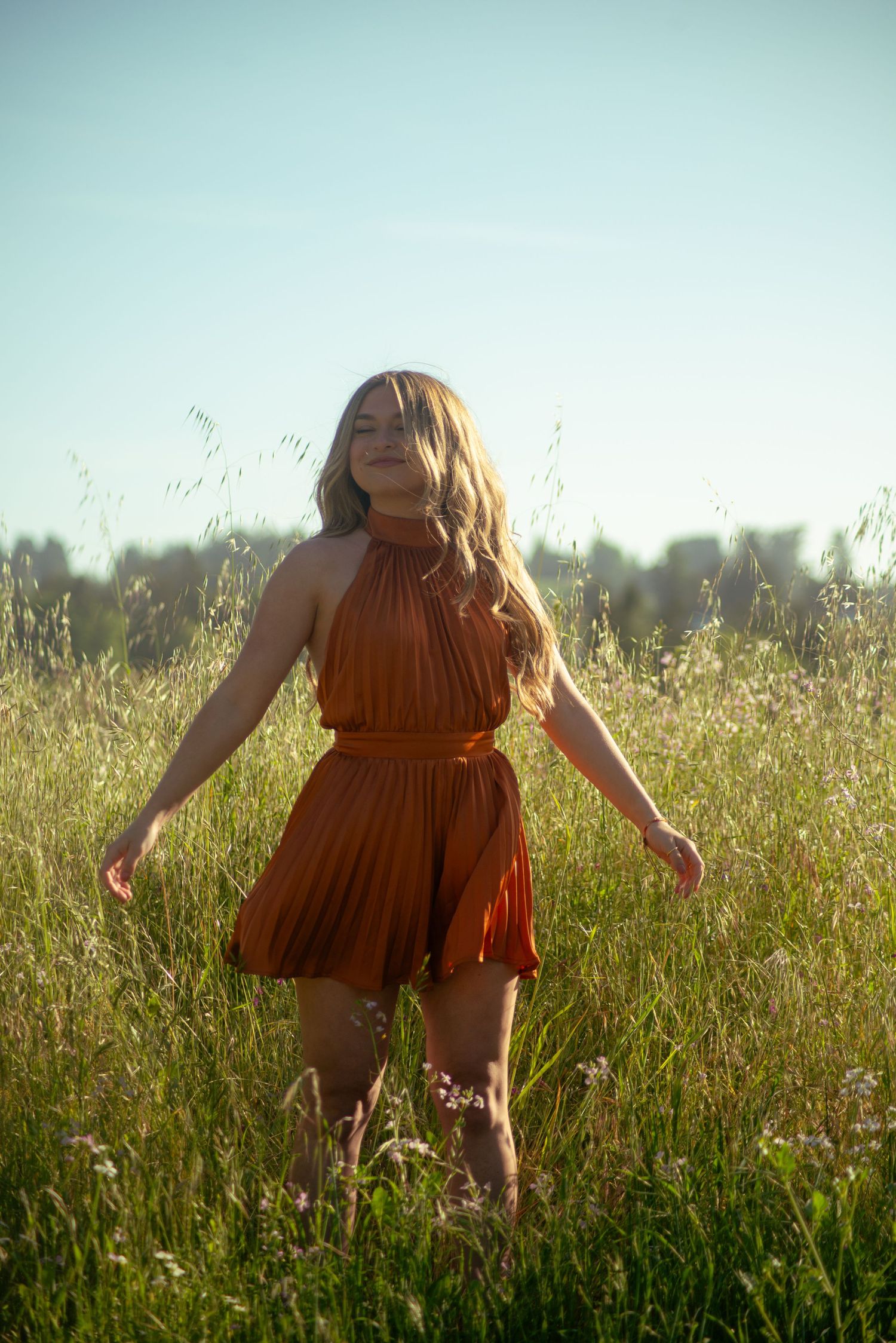 Someone in a flowing rust dress twirls playfully in a sunlit meadow with tall grass.
