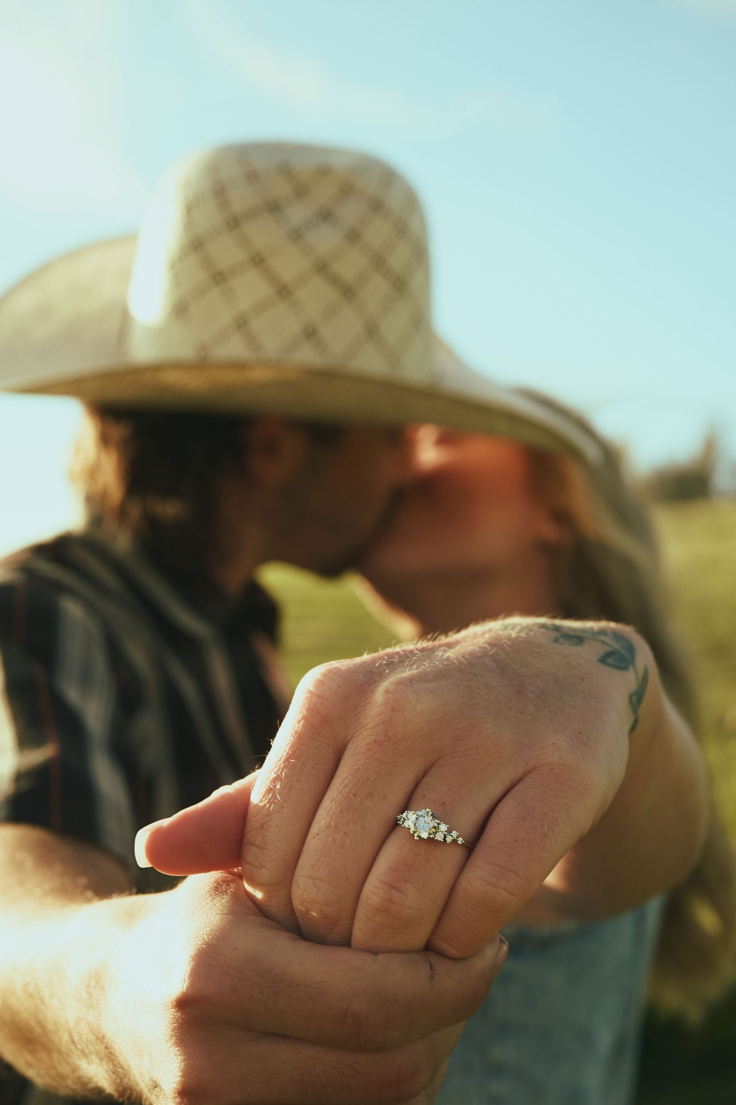 Couple kissing outdoors displaying diamond engagement ring, woman wearing straw cowboy hat in sunny field.