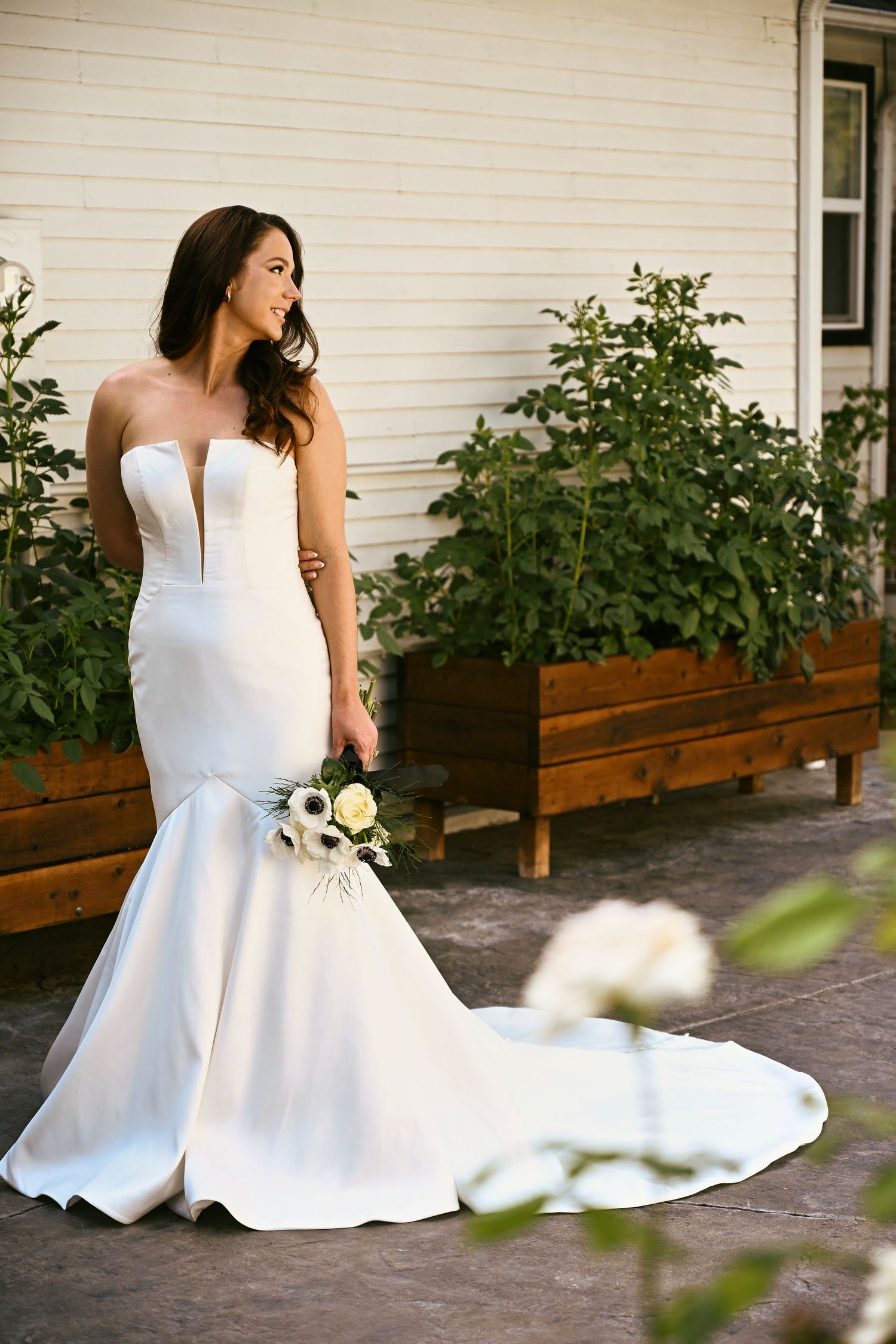 Bride in strapless mermaid wedding gown holding white bouquet, posing outdoors near garden planters.