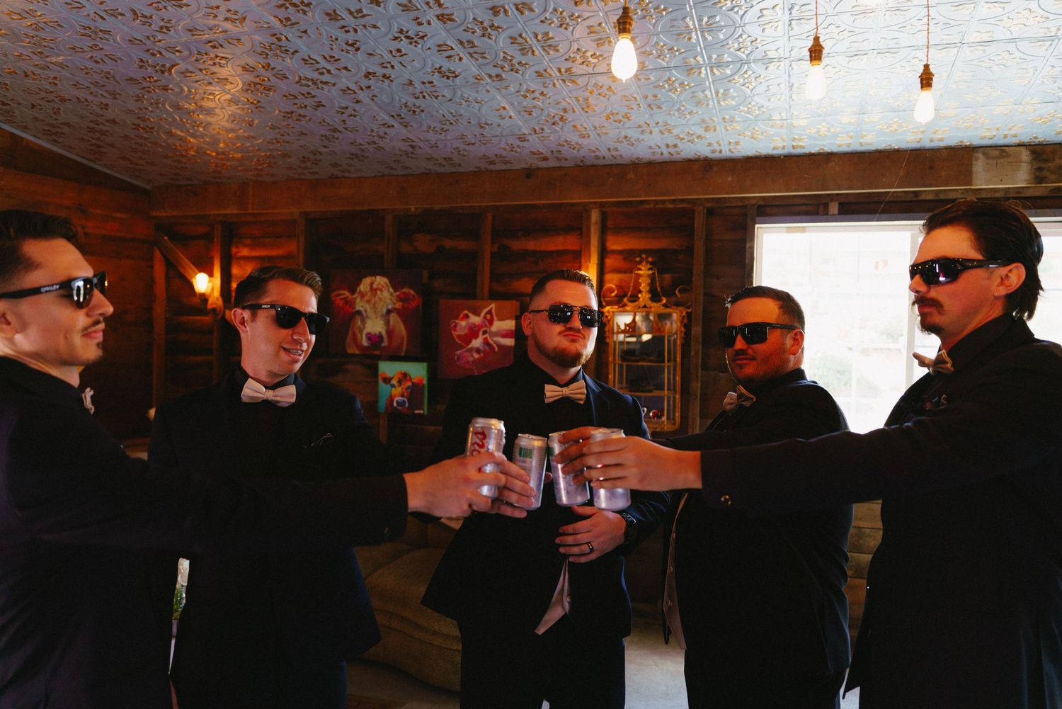 Five groomsmen in black tuxedos and sunglasses toasting with drinks inside a rustic venue.