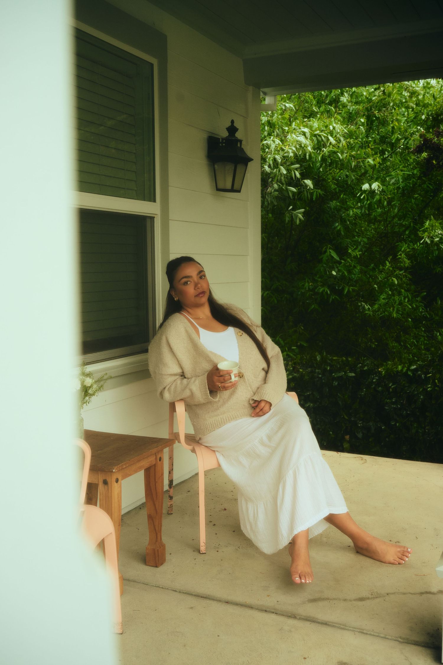 Woman in white dress and beige cardigan sitting barefoot on porch holding a cup.