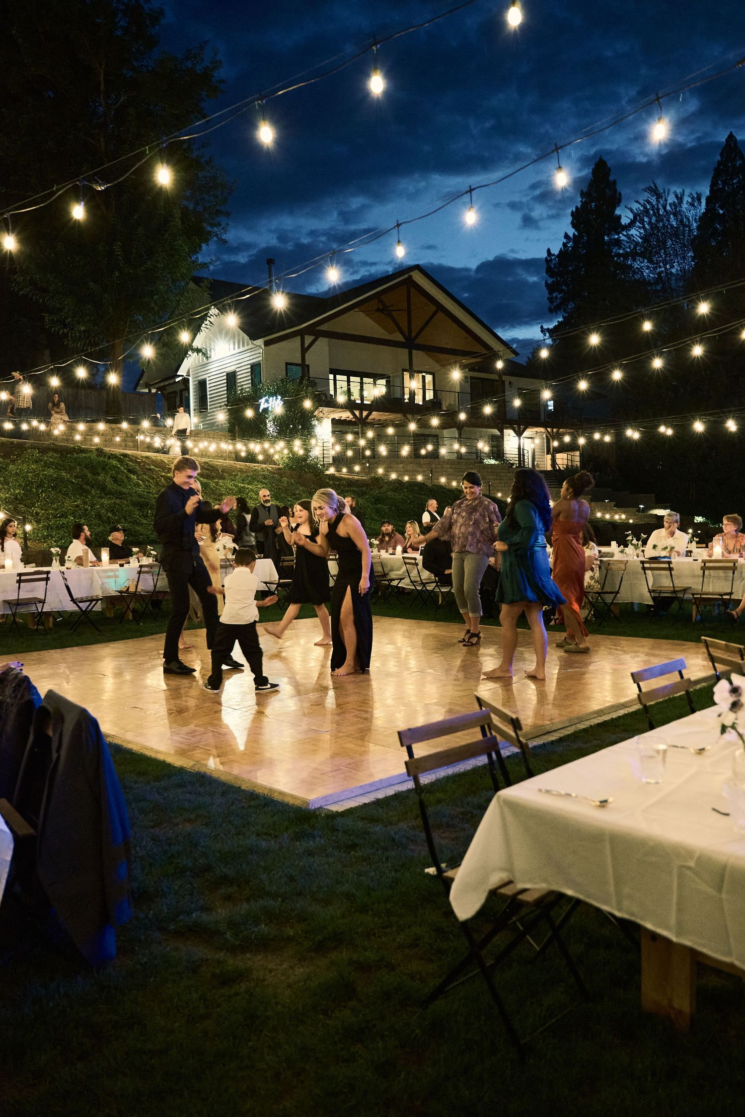 Outdoor evening wedding reception with string lights, guests dancing on a wooden floor, surrounded by tables and a house backdrop.