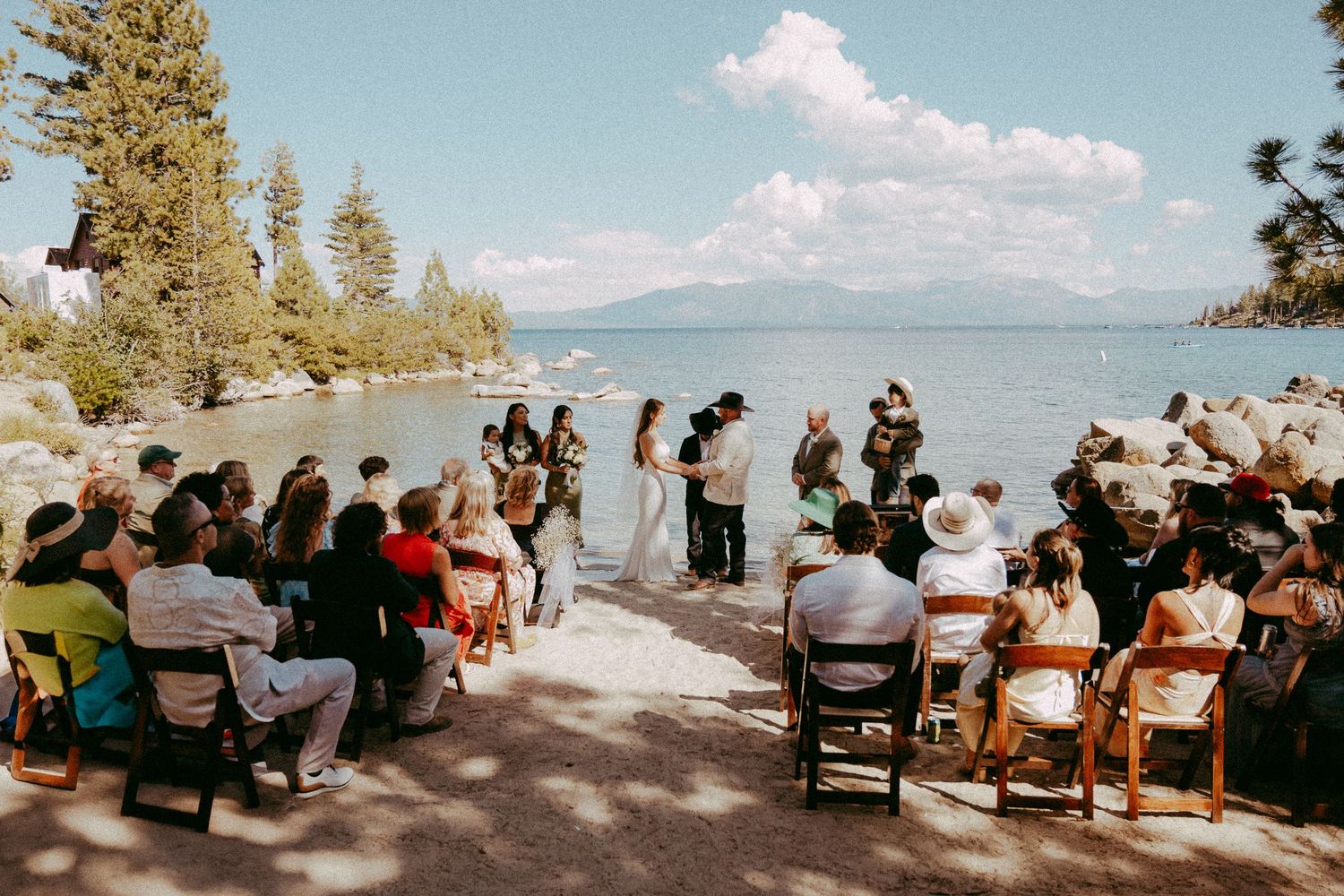 Outdoor lakeside wedding ceremony with guests seated on wooden chairs, couple at altar, Lake Tahoe backdrop.
