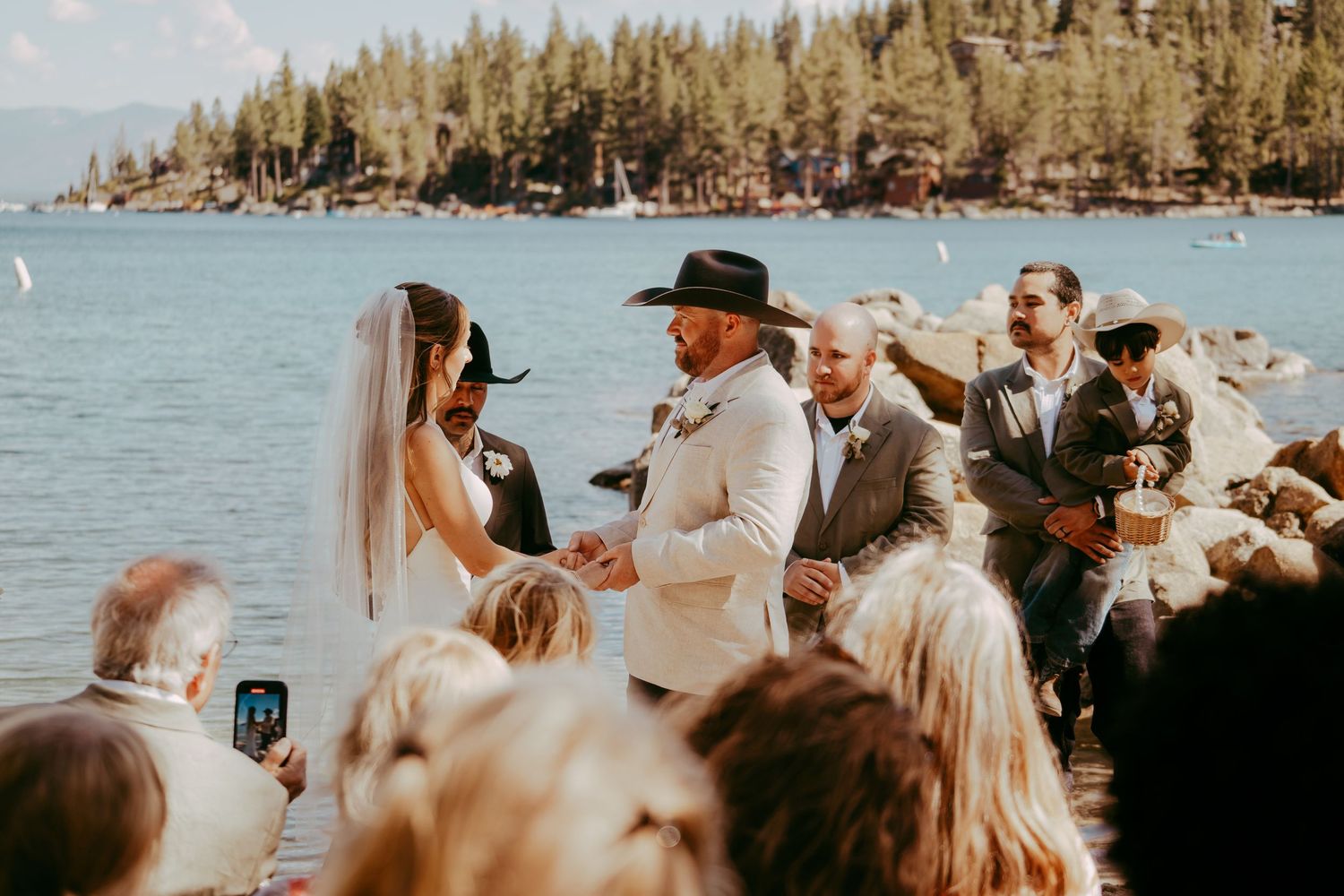 Outdoor lakeside wedding ceremony with bride and groom exchanging vows, pine trees and blue water in background.