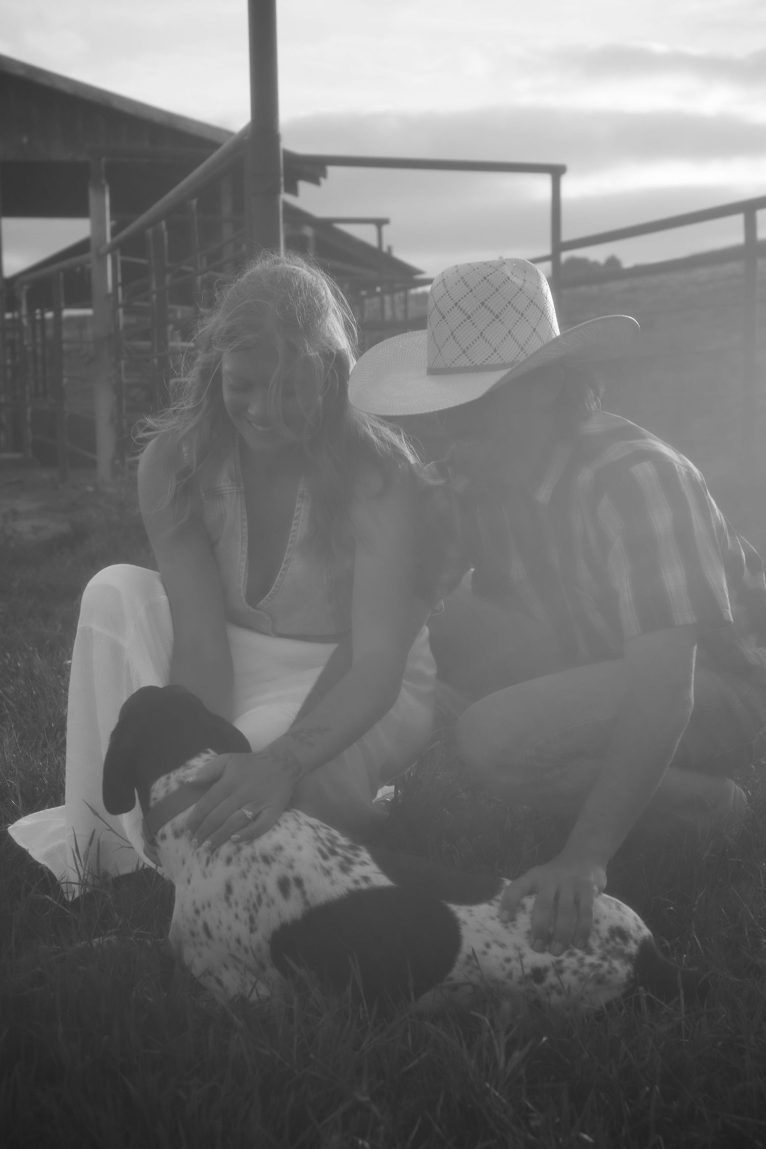 Black and white photo of a couple crouching on grass petting a spotted dog near a farm fence at sunset.