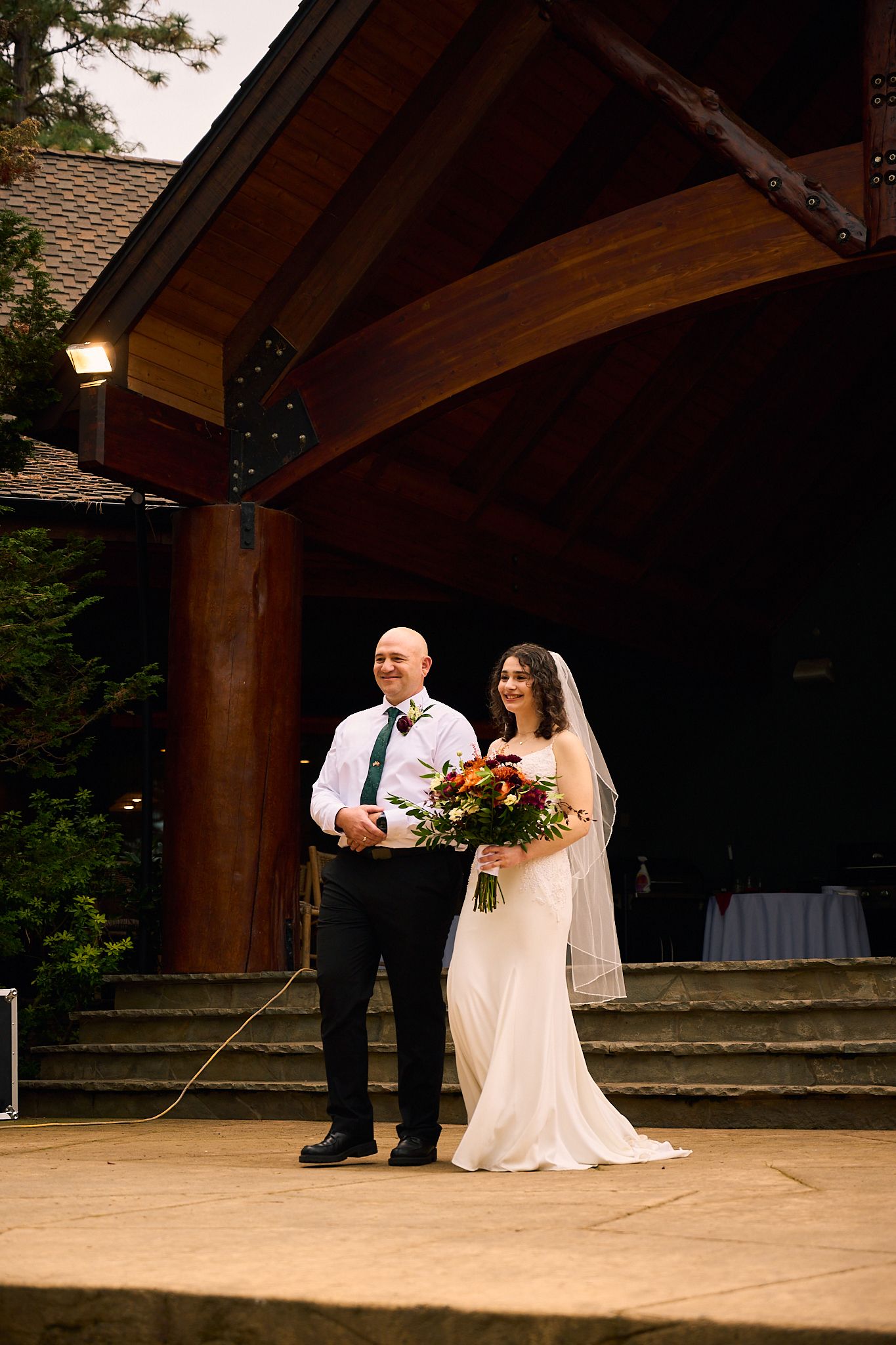 Bride in white gown with veil holding bouquet stands with groom in black suit outside rustic wooden wedding venue.