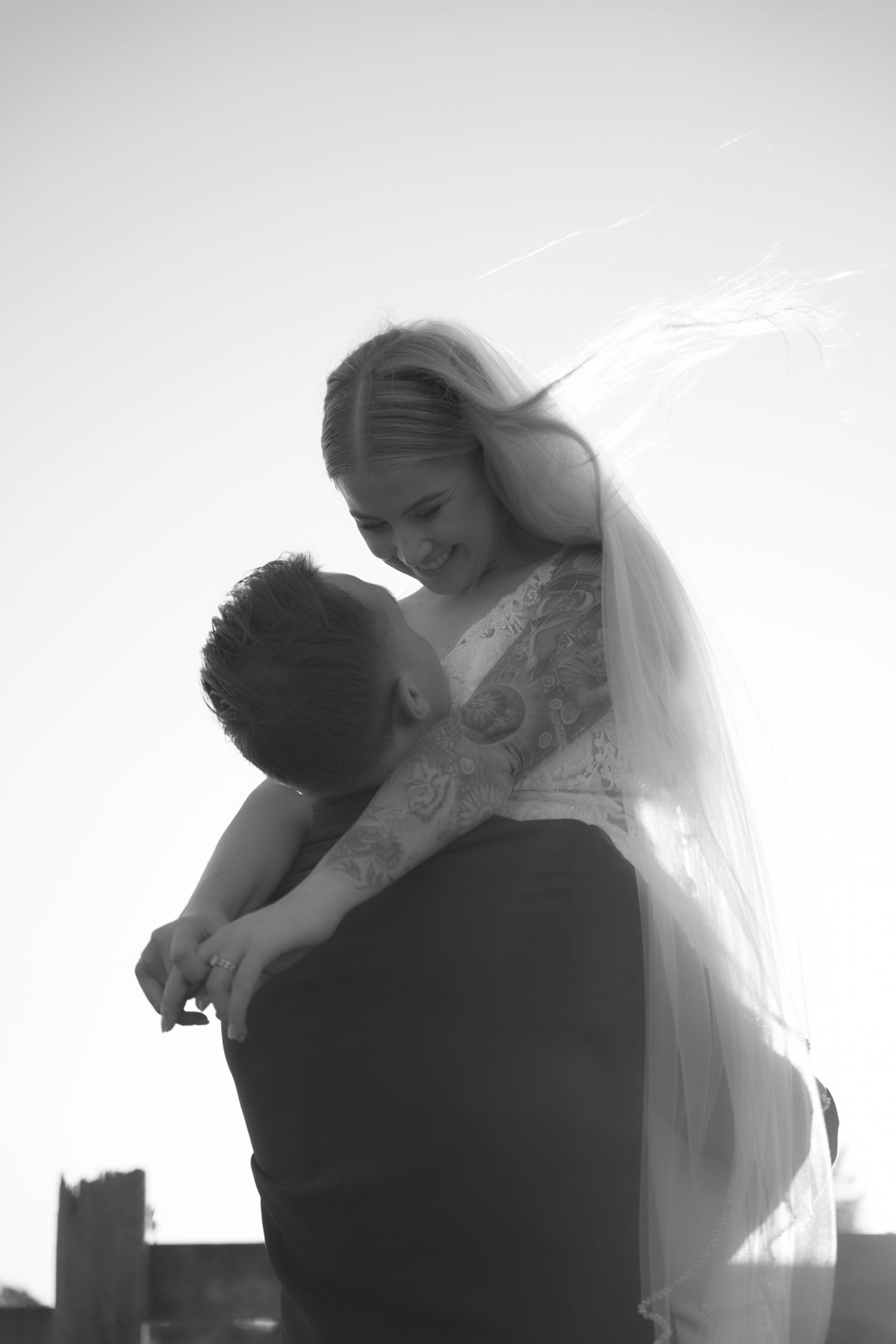 Black and white photo of groom lifting bride in wedding dress and veil against bright sky.