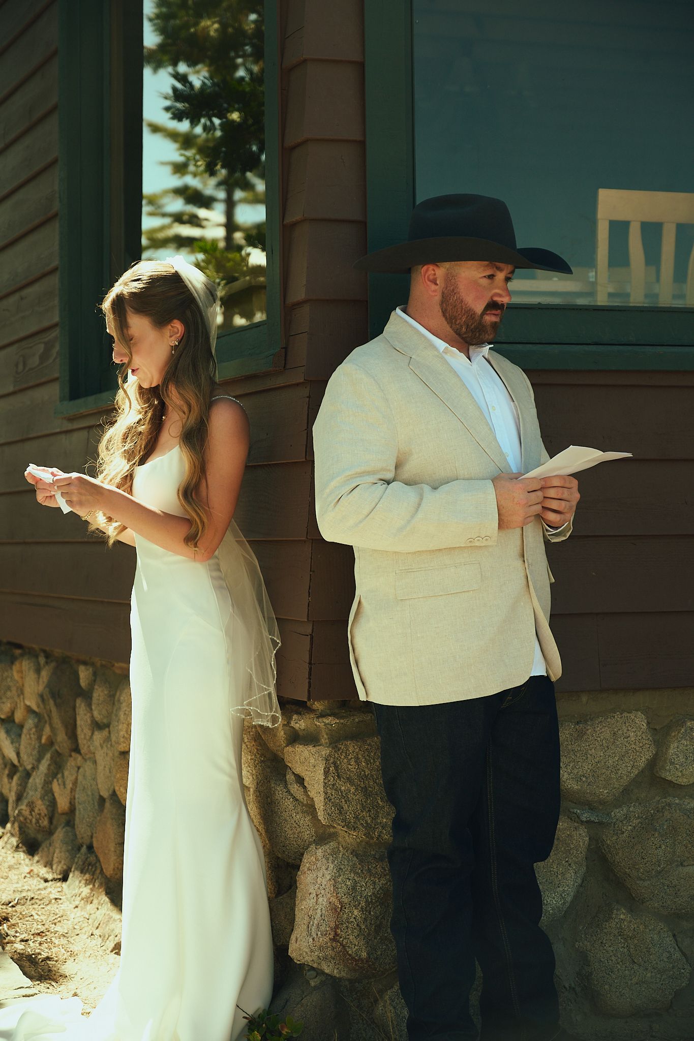 Bride in white gown and groom in tan blazer and cowboy hat read vows separately before wedding ceremony outdoors.