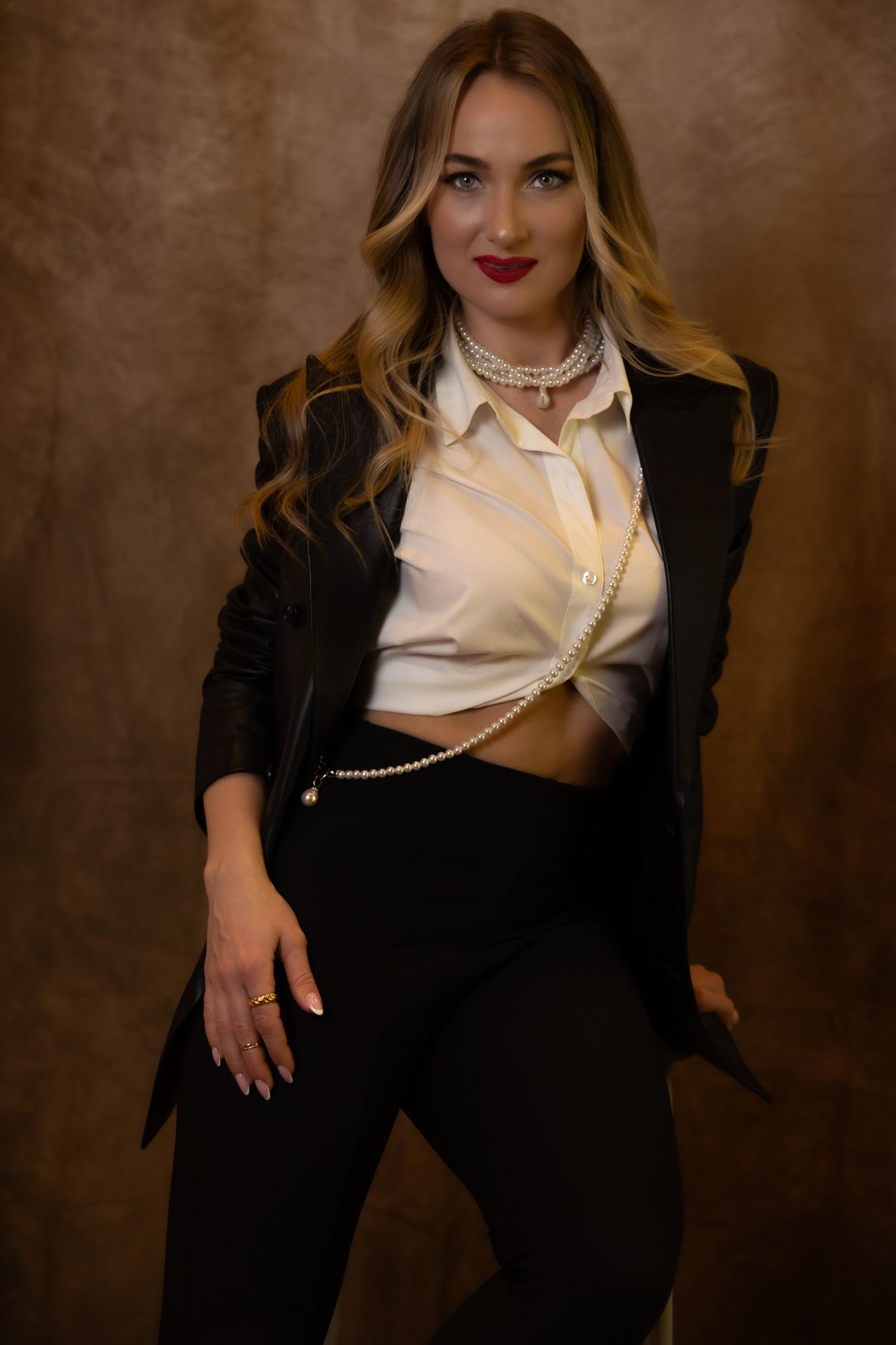 Fashion model in black suit and white crop top with pearl accessories against brown background.