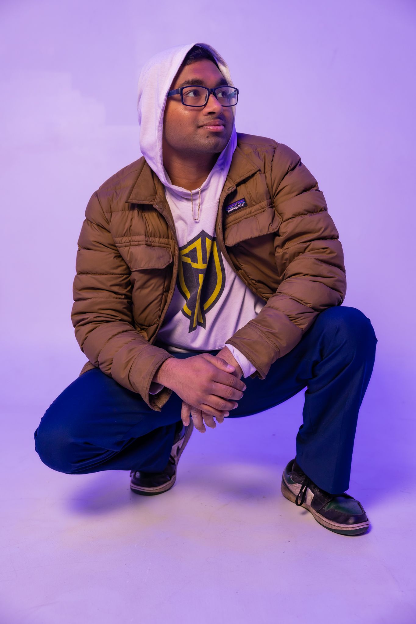 Casual portrait in brown puffer jacket and white hoodie crouching against purple backdrop.